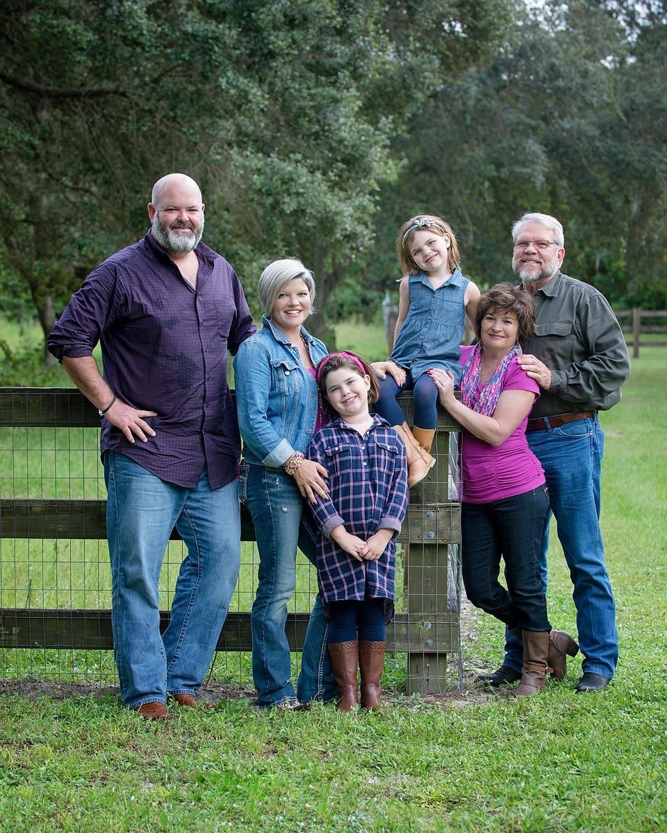 Family portrait: adults and children standing near a wooden fence in a green outdoor setting.