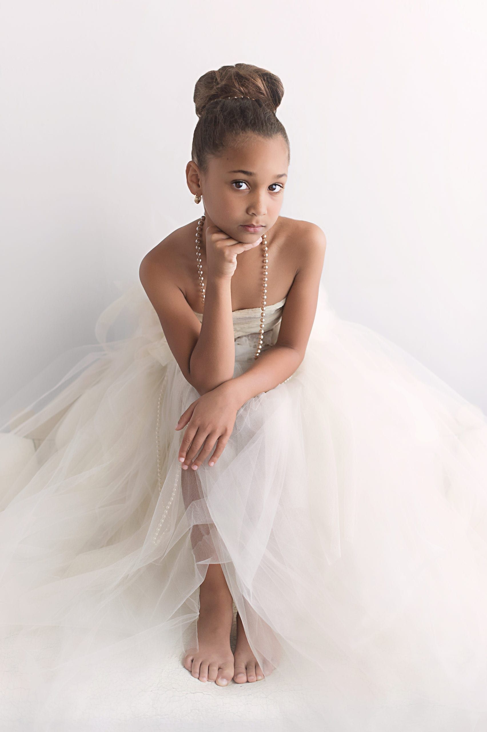 A young girl in a white dress sits thoughtfully, chin on hand, with hair up.