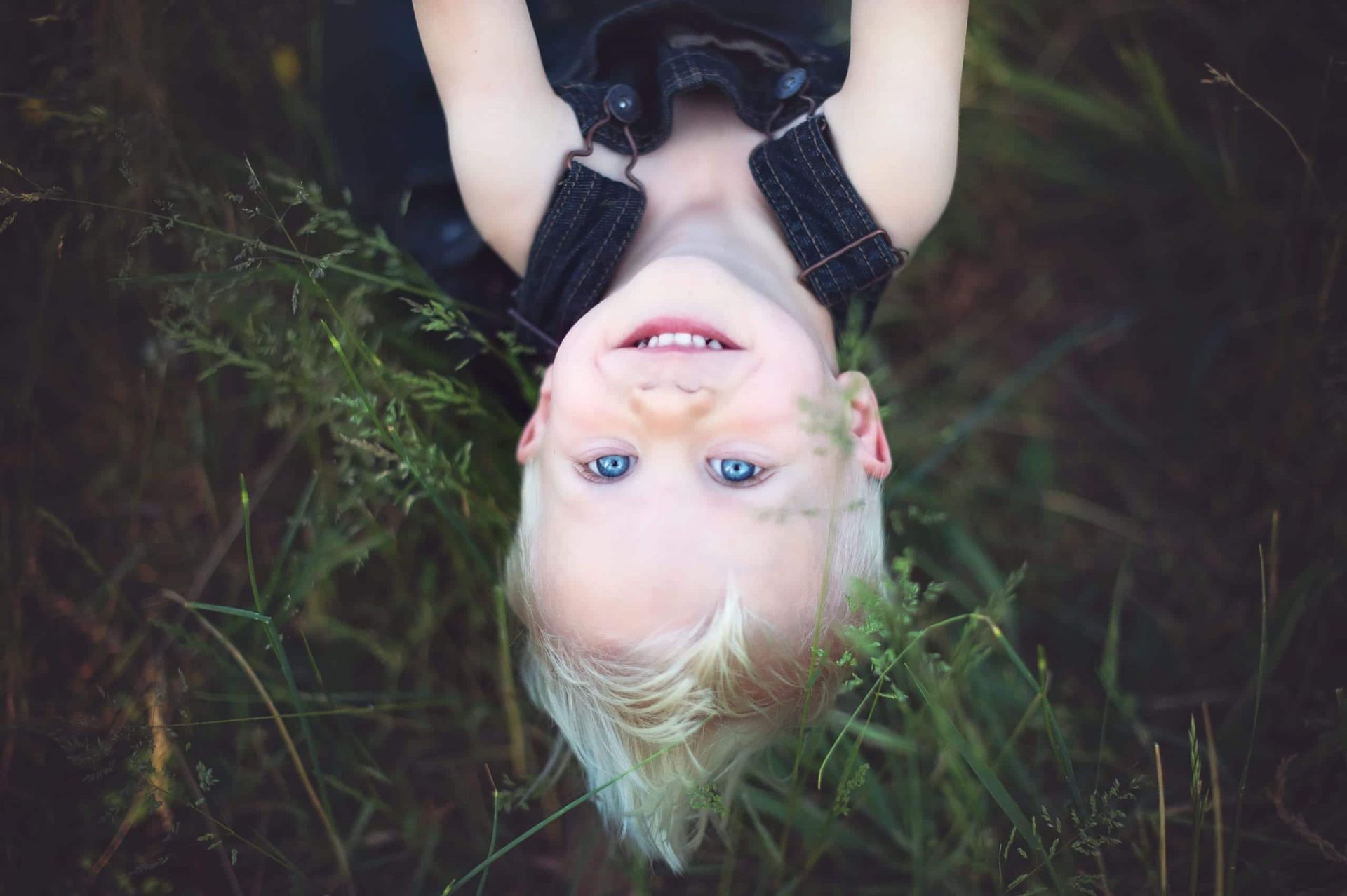 Blond-haired child with blue eyes smiling upside down in tall green grass, wearing black overalls.