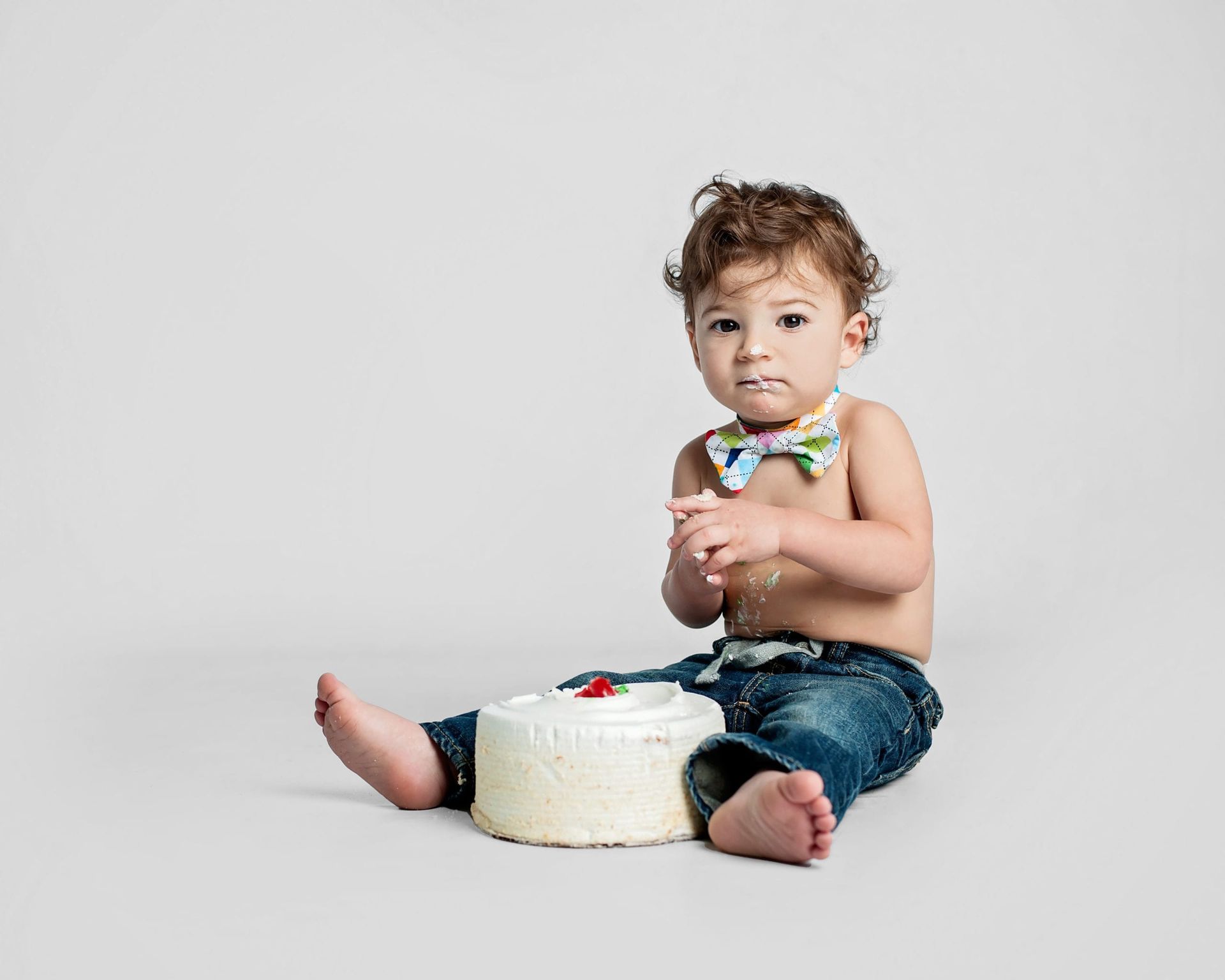 A baby with a bow tie sits next to a cake, covered in frosting.