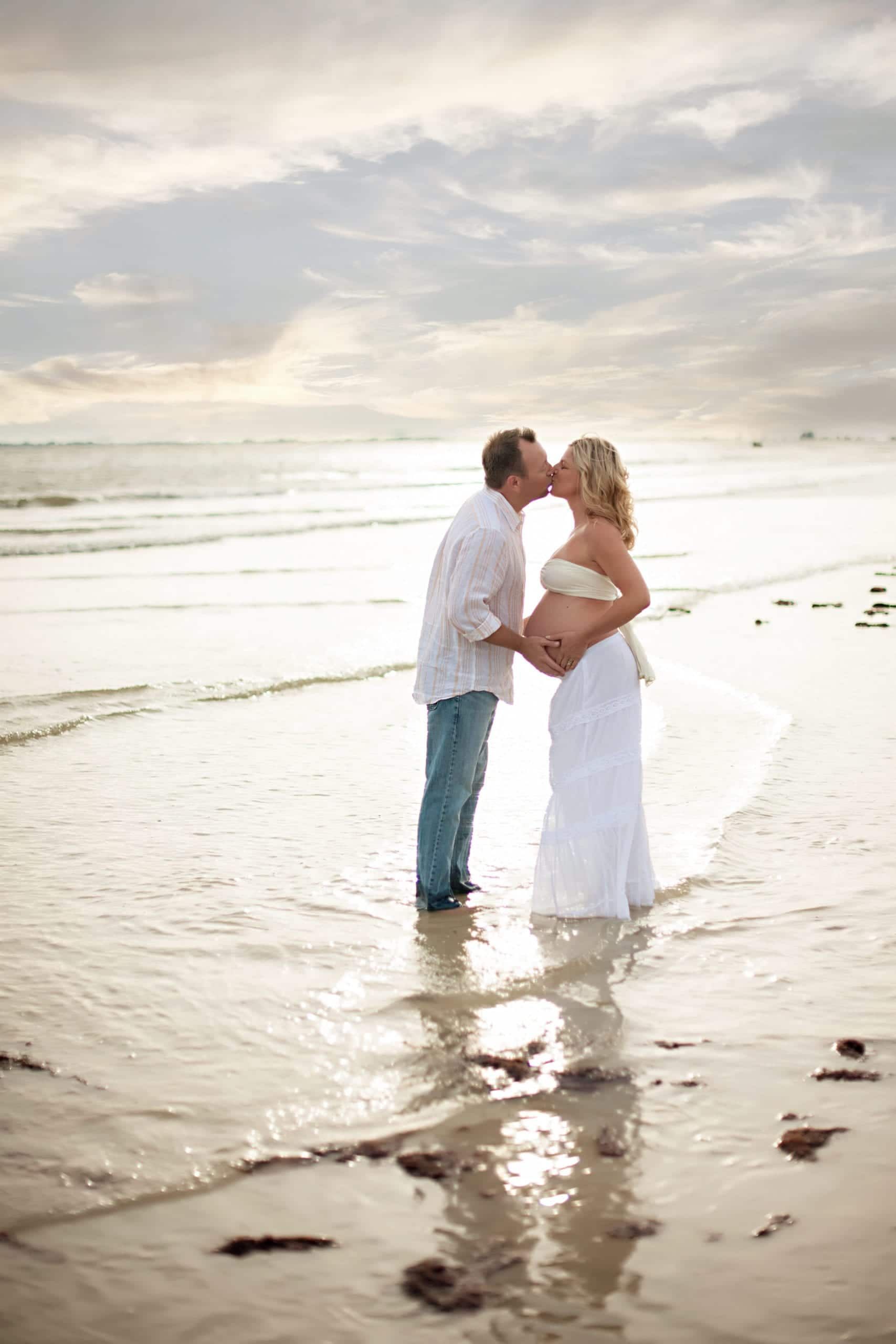 Pregnant woman and man kissing on a beach at sunset. She wears a white dress, he wears a white shirt and jeans.