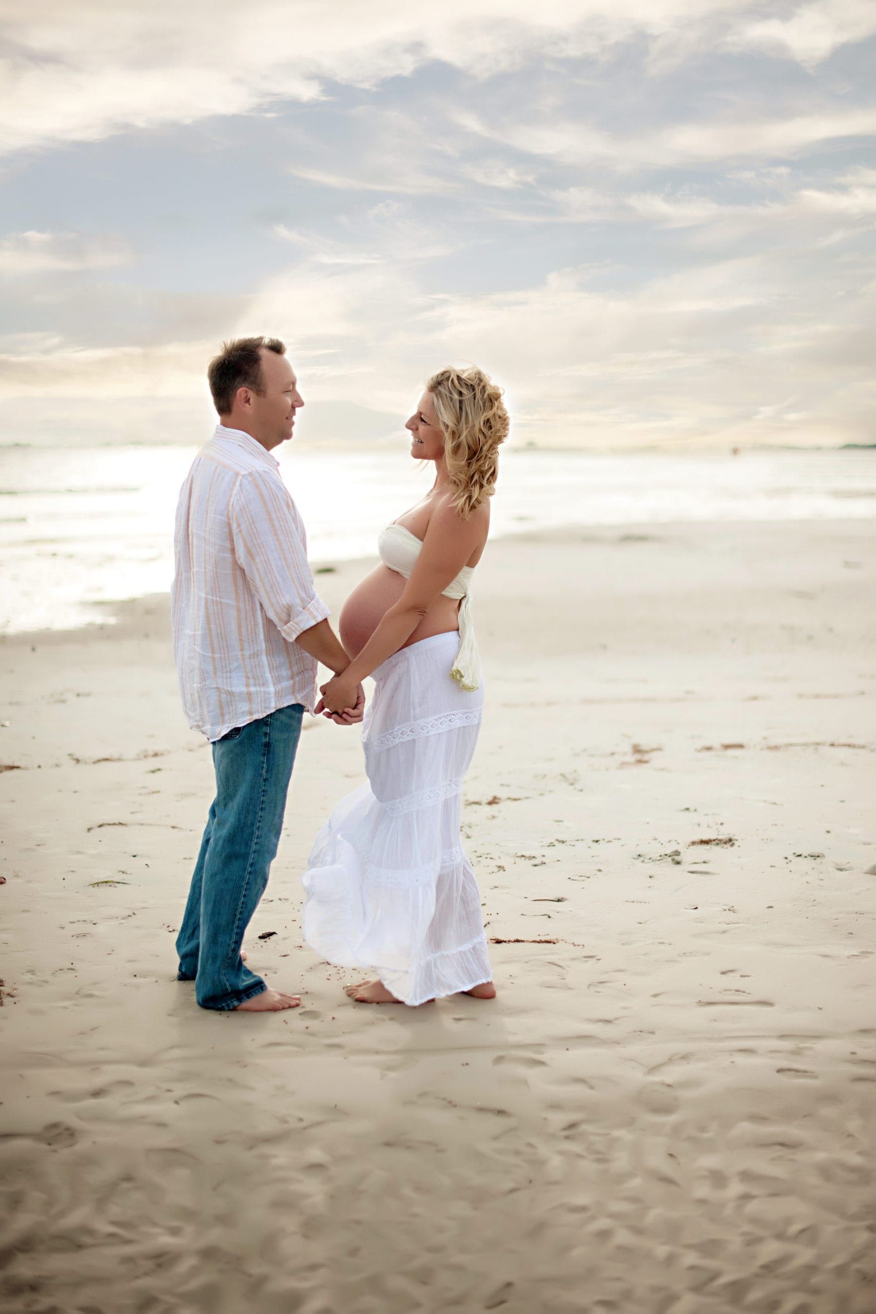 Pregnant couple holding hands, looking at each other on a beach. Sky is cloudy. She wears white, he wears blue jeans and a white shirt.