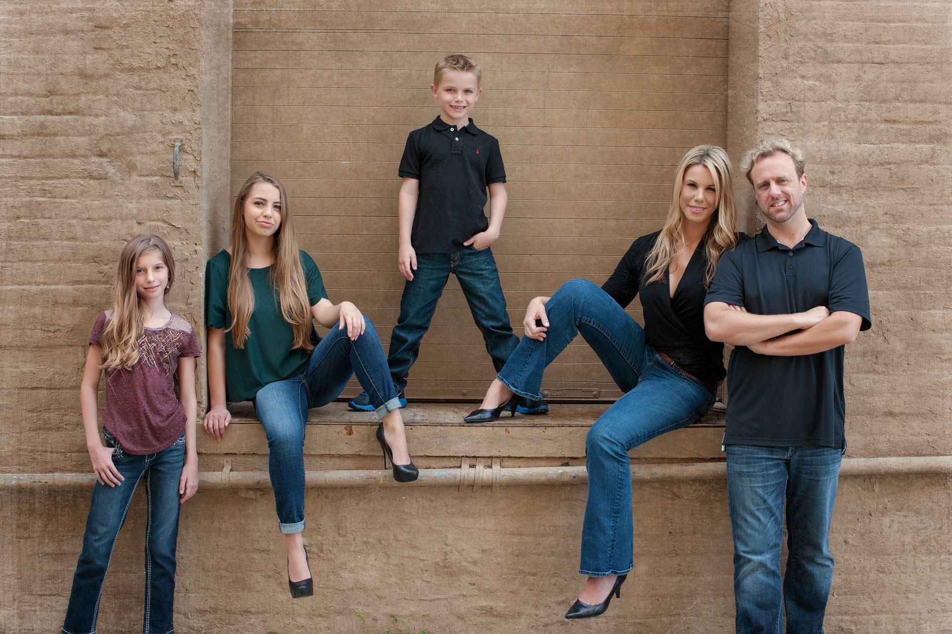 Family of five poses outdoors against a textured wall. Adults and teens wear jeans and shirts. Boy stands in the middle.