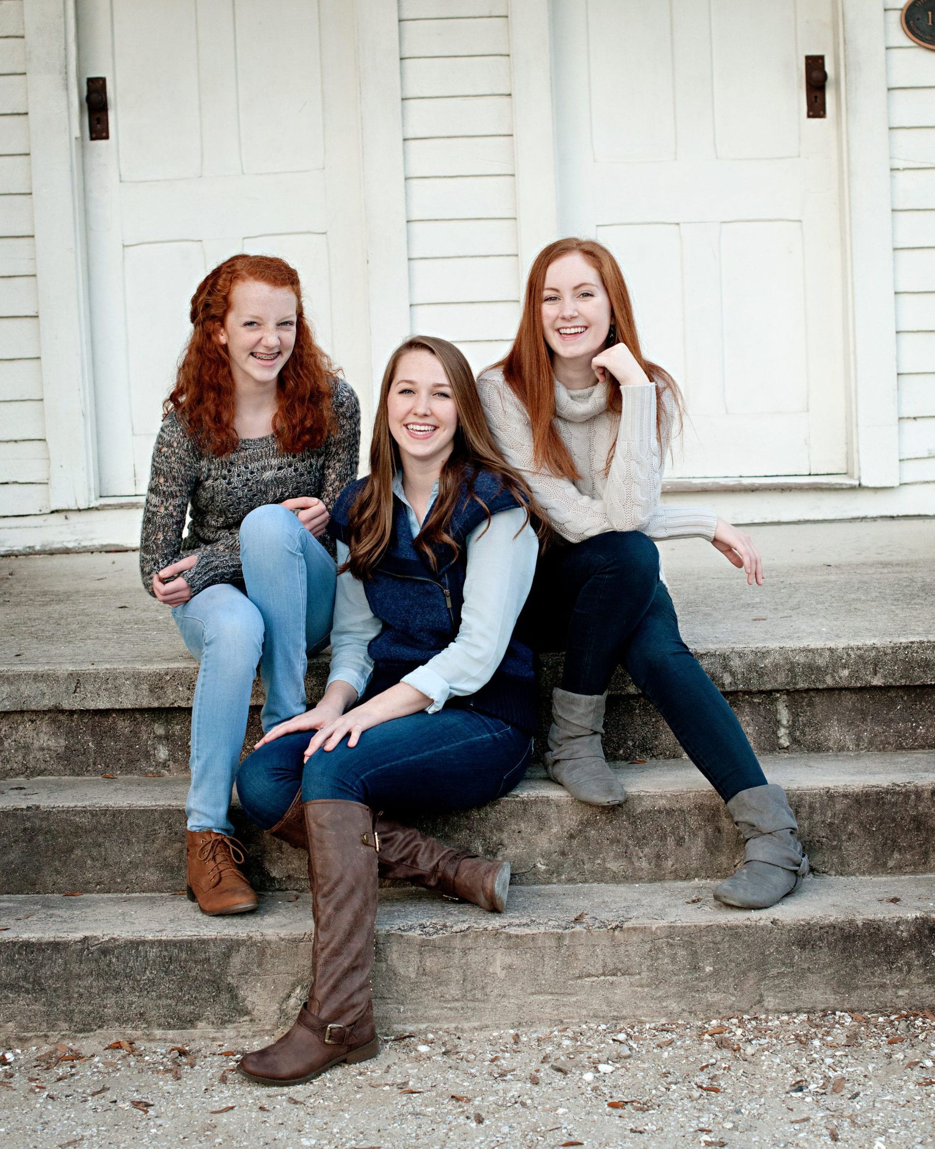 Three red-haired young women sit on steps. Building with white doors in background.