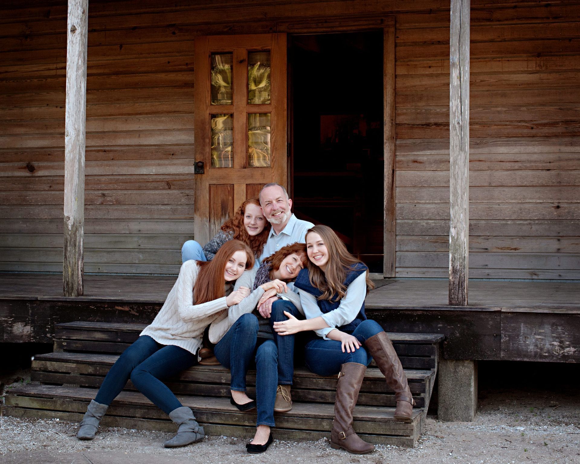 Family of five smiling, embracing on wooden porch steps. Father, three daughters. Rustic cabin background.