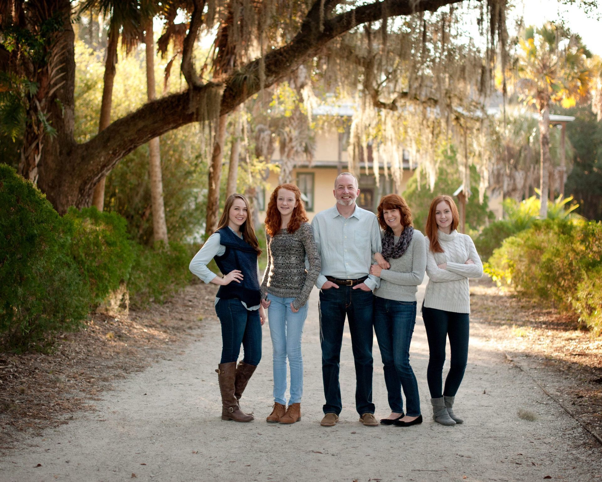 Family of five poses on a sandy path, under a large tree with hanging moss, in front of a house.