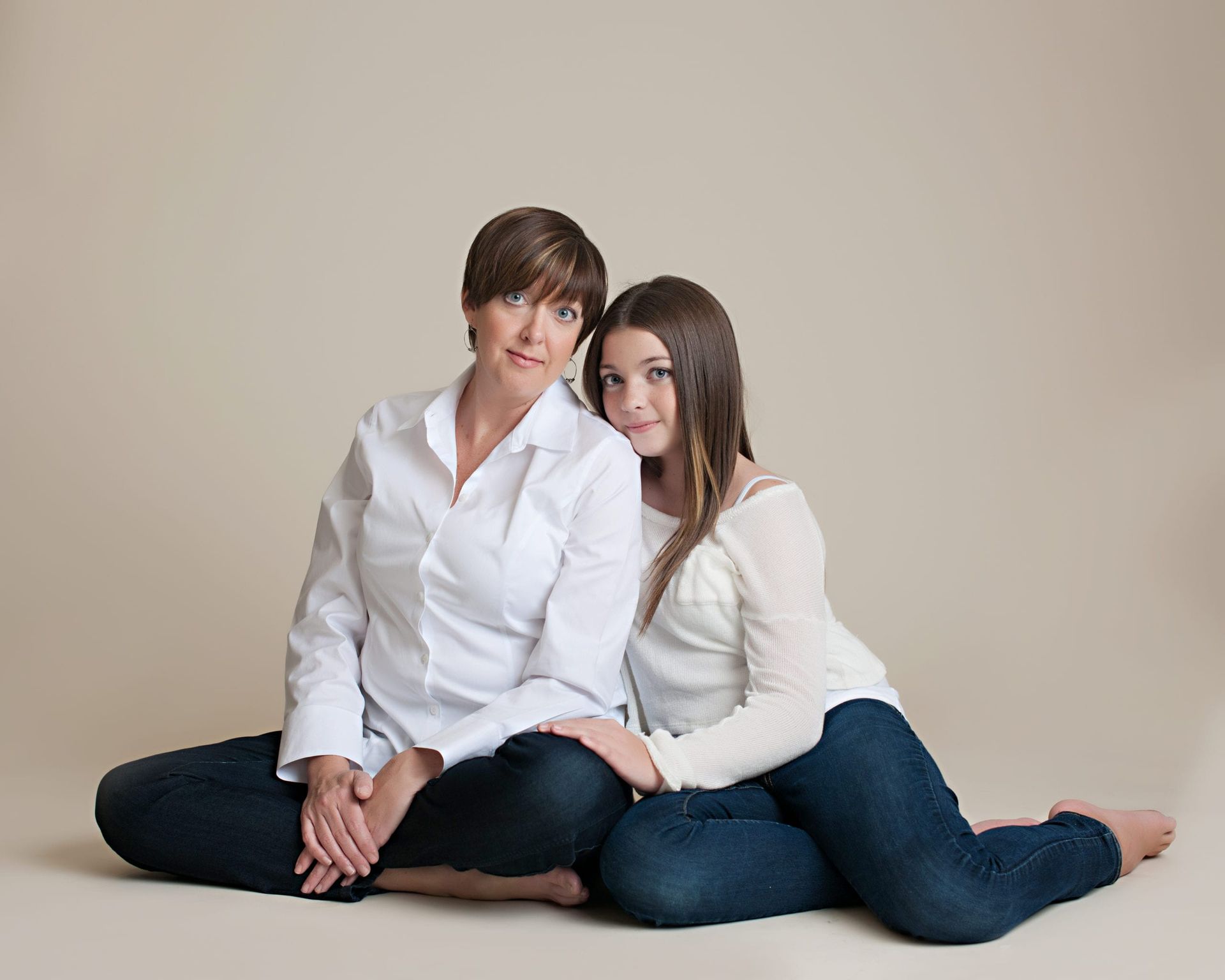 Woman and girl sitting together on floor, wearing white shirts and jeans, smiling.
