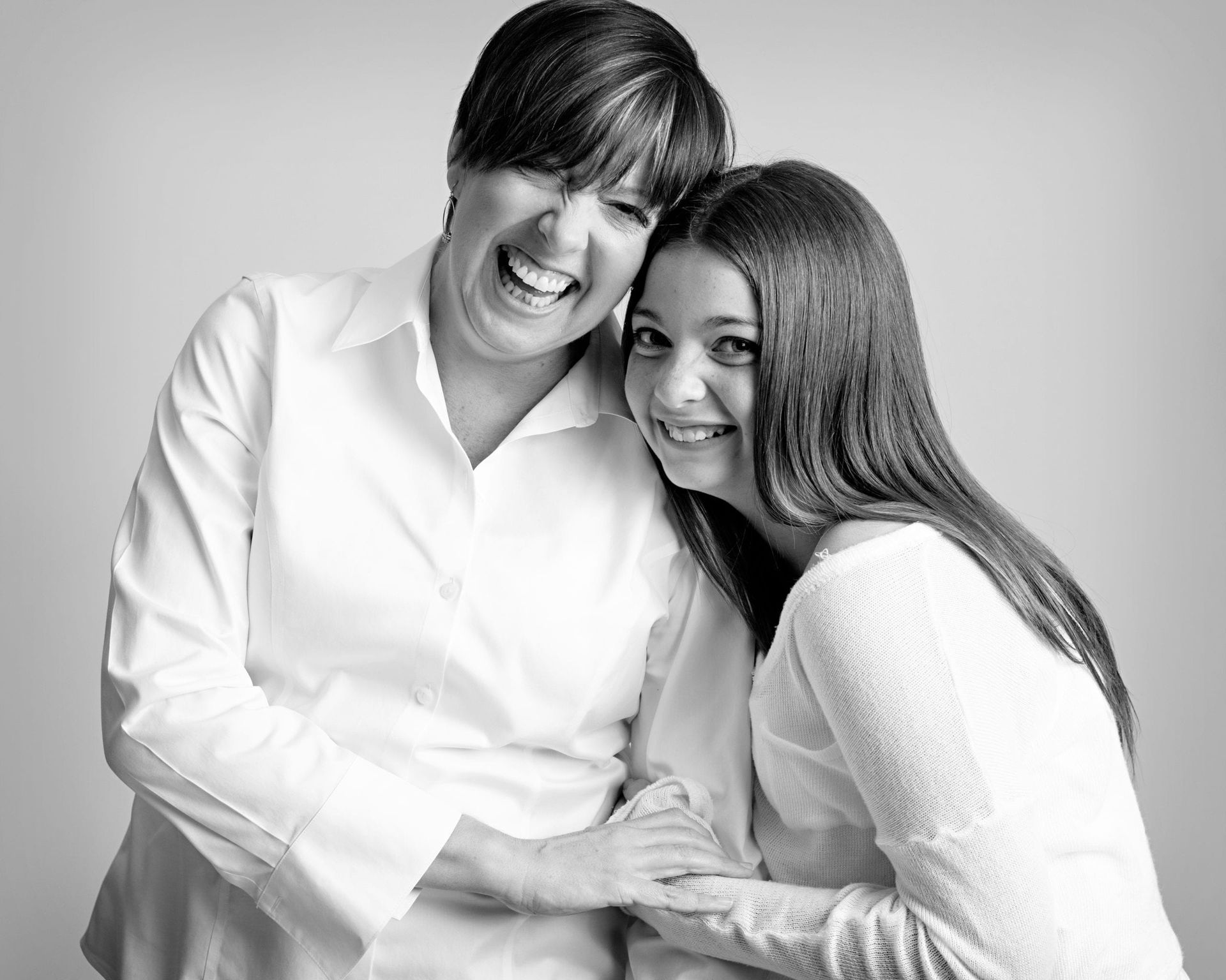 Woman and teen girl smiling, embracing in front of a light gray background.