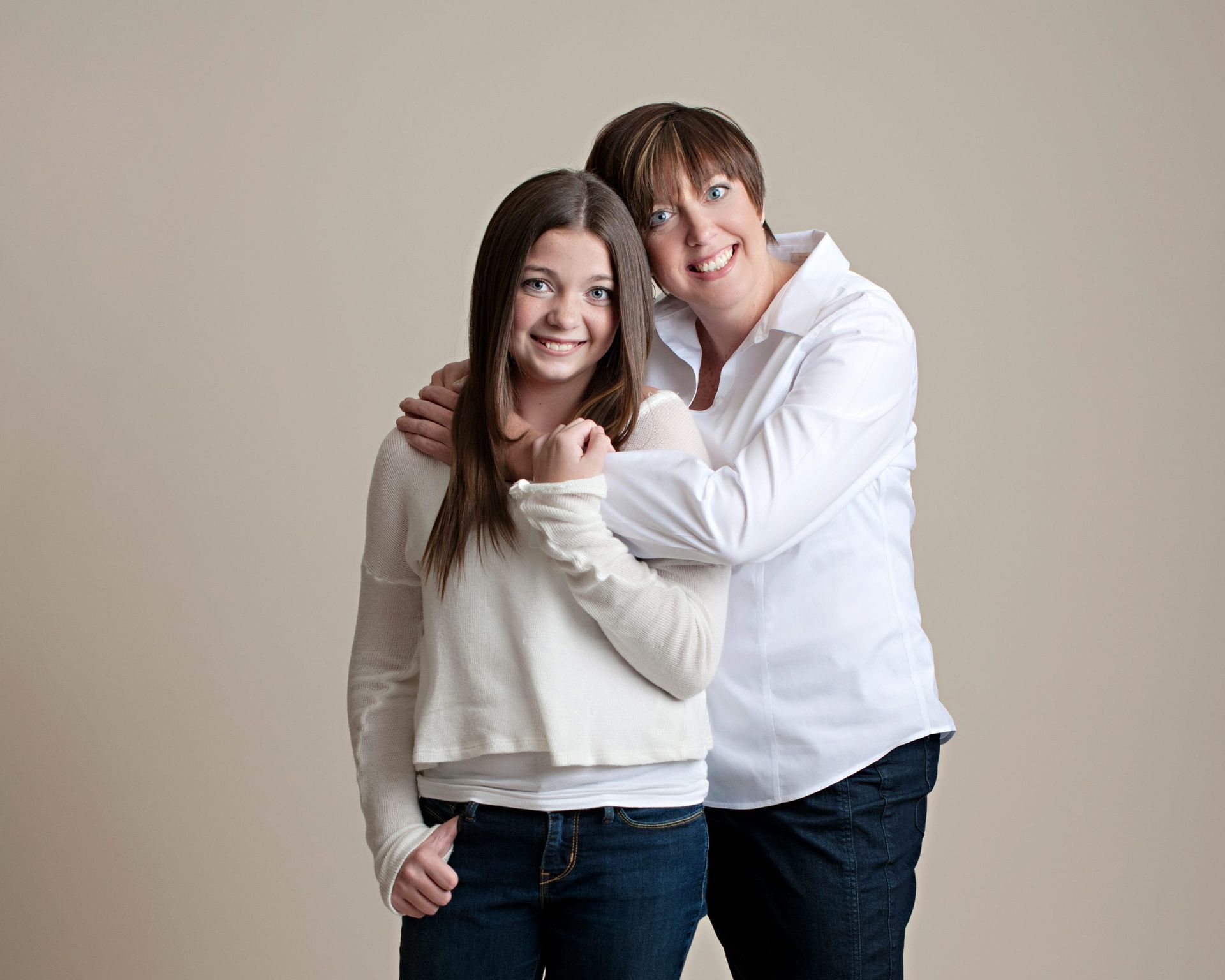 Mother and daughter, smiling, arm around each other. White tops, blue jeans, neutral background.