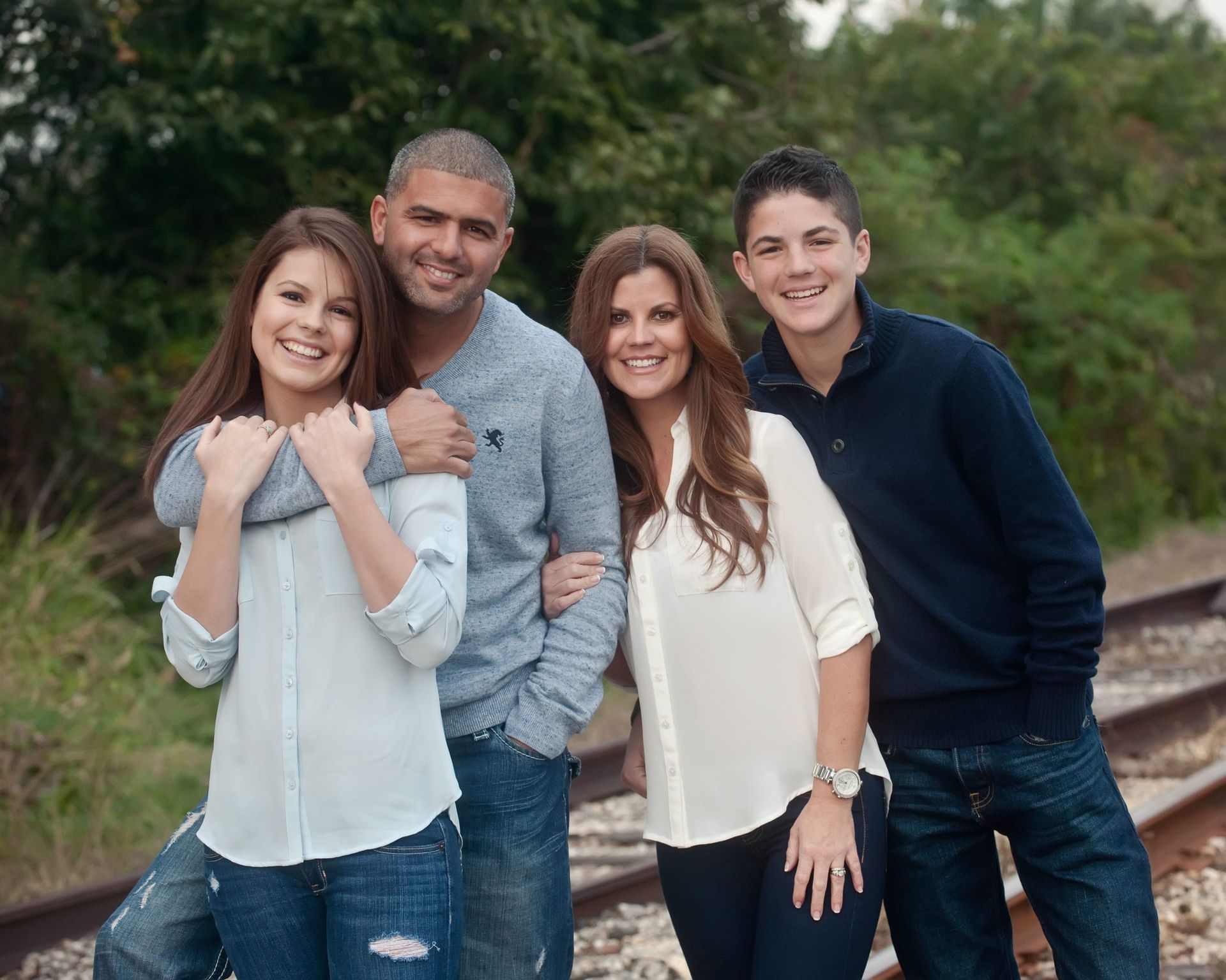 Family of four smiling near train tracks. Adults embrace while teens stand beside them.