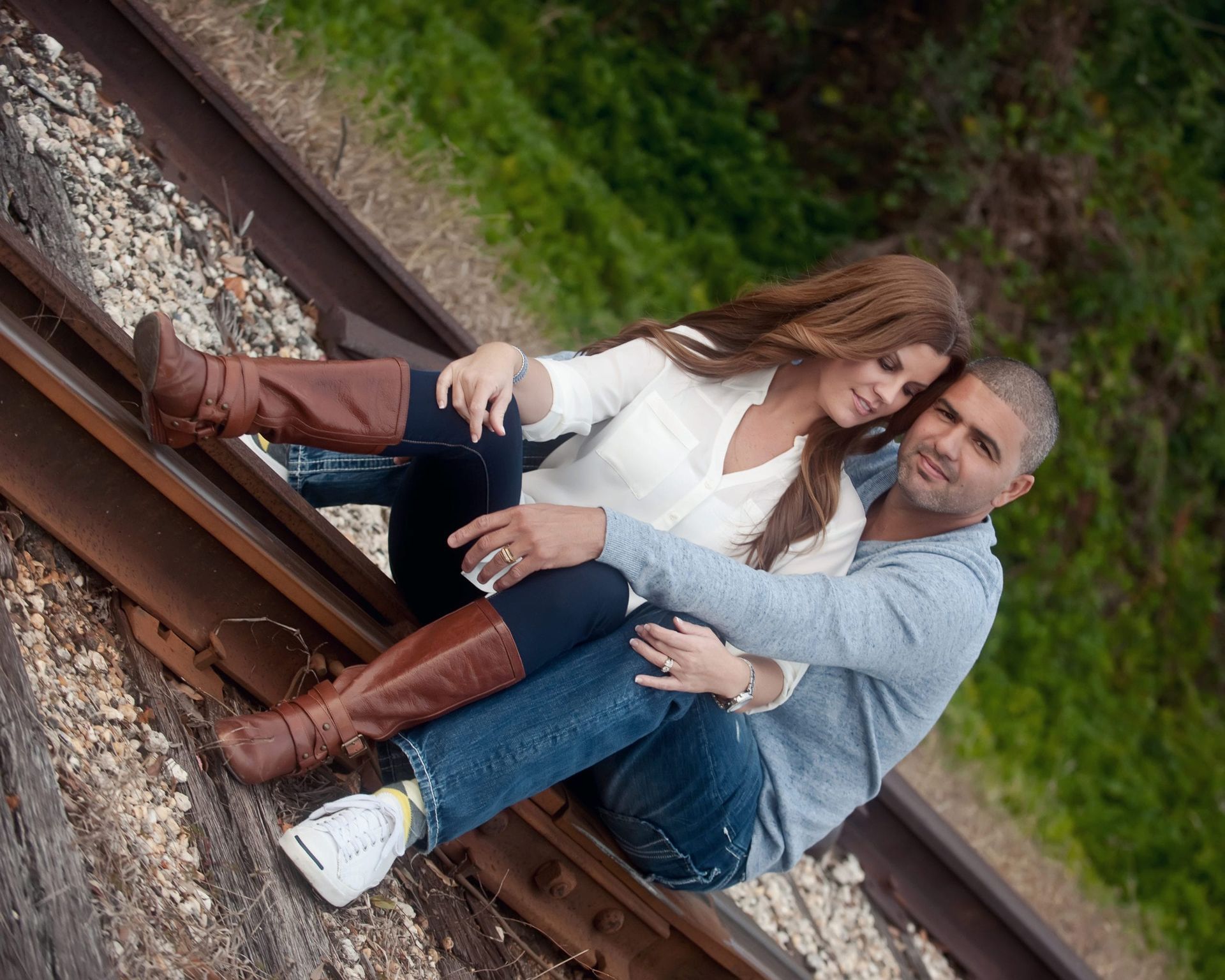 Couple embraces on a railroad track. Man in blue, woman in white and brown boots. Green backdrop.