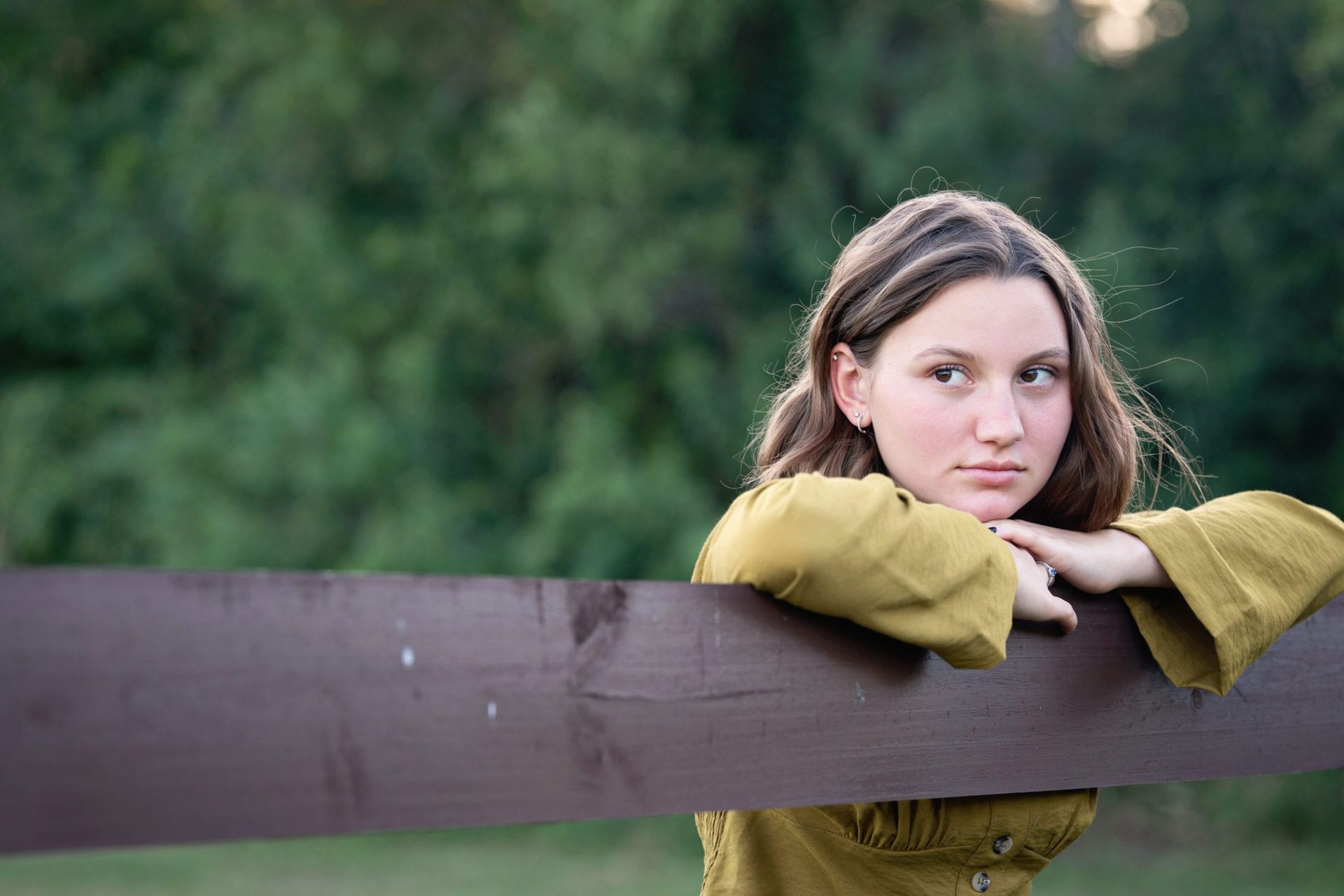 Teenage girl with brown hair, leaning on a brown fence, looking thoughtful in a green outdoor setting.