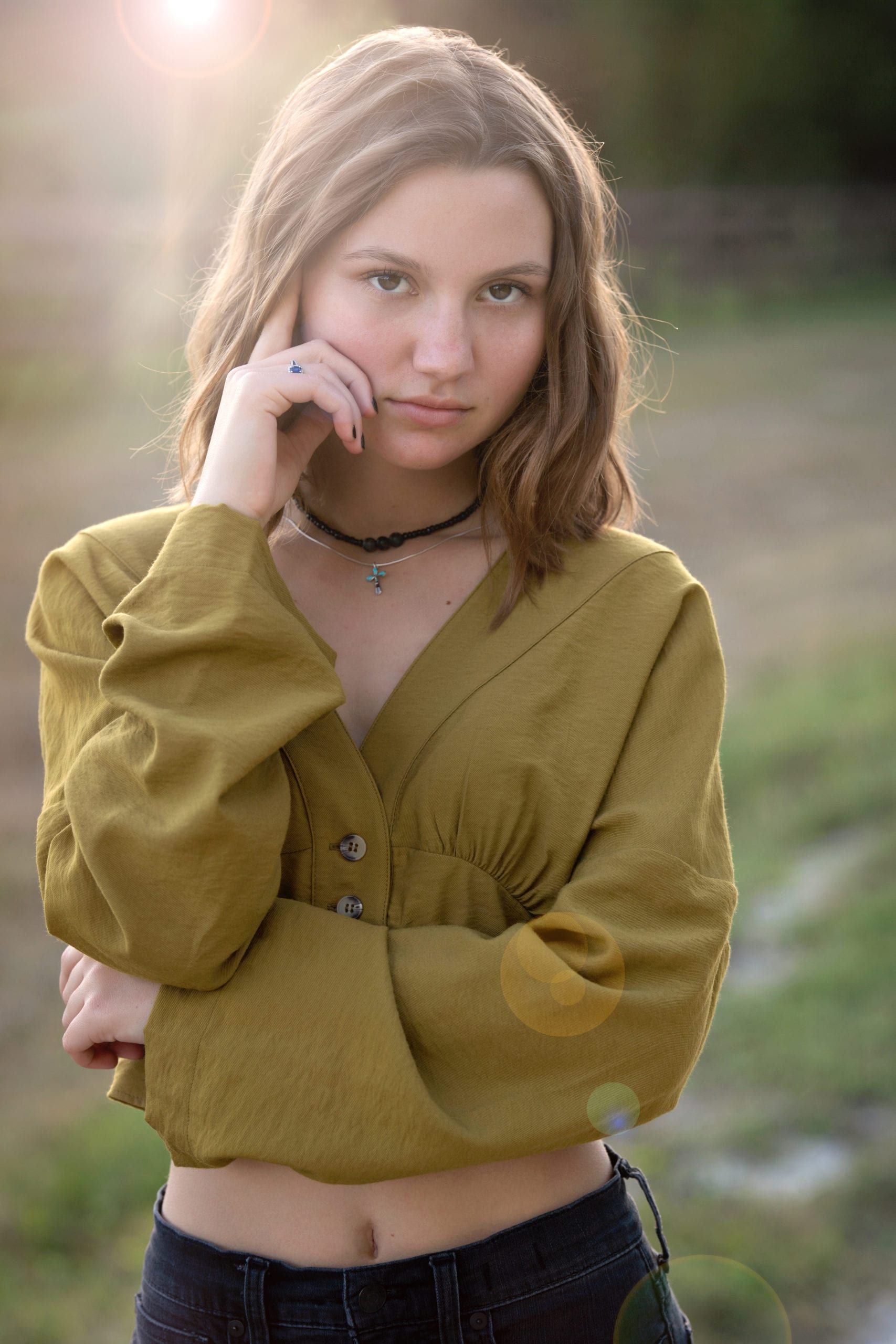 Woman with light brown hair, wearing a gold top, looking pensively at the camera outdoors with a blurred background.