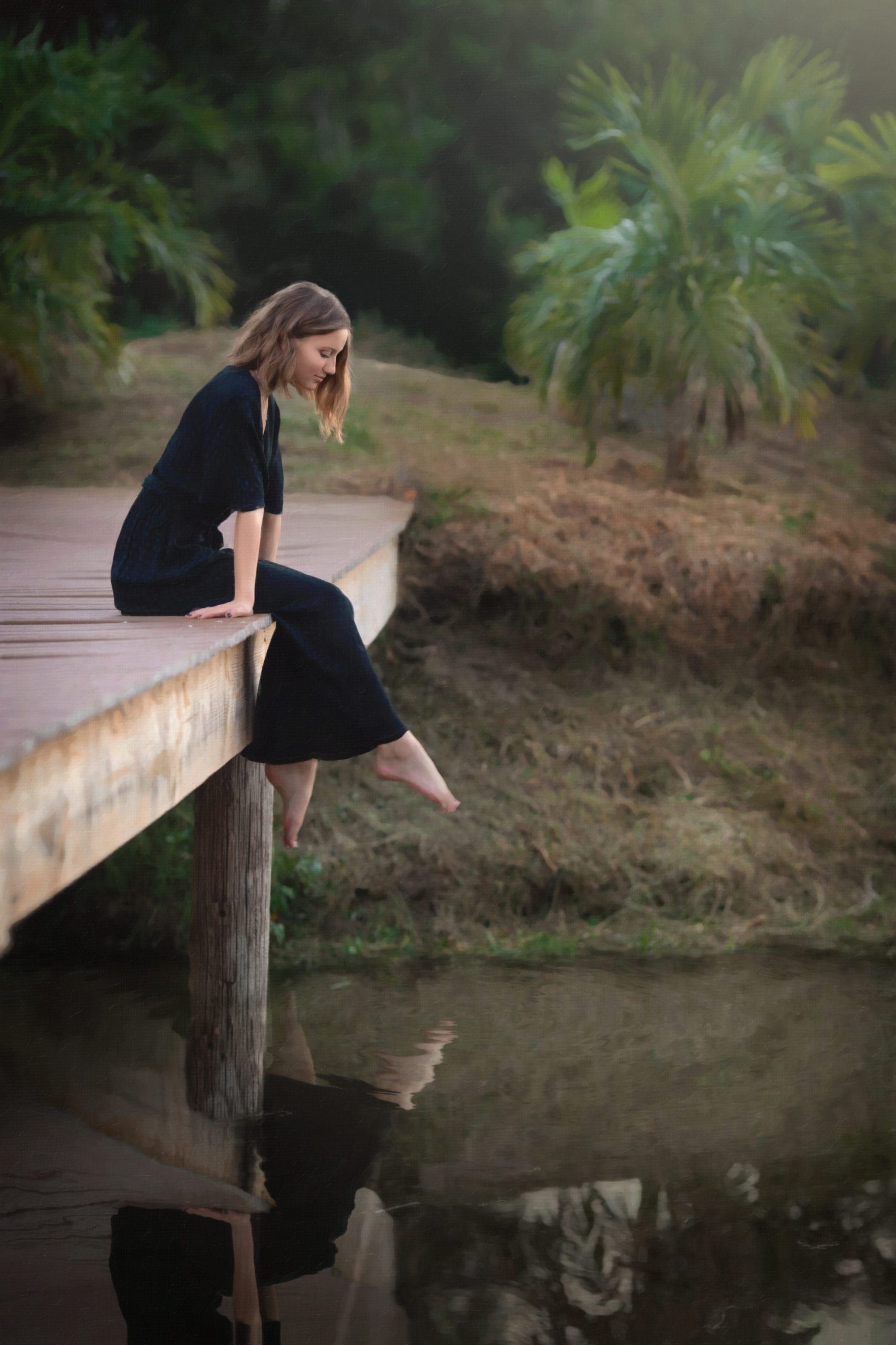 Woman in black dress sits on dock, dipping feet into water, looking down. Tropical setting.