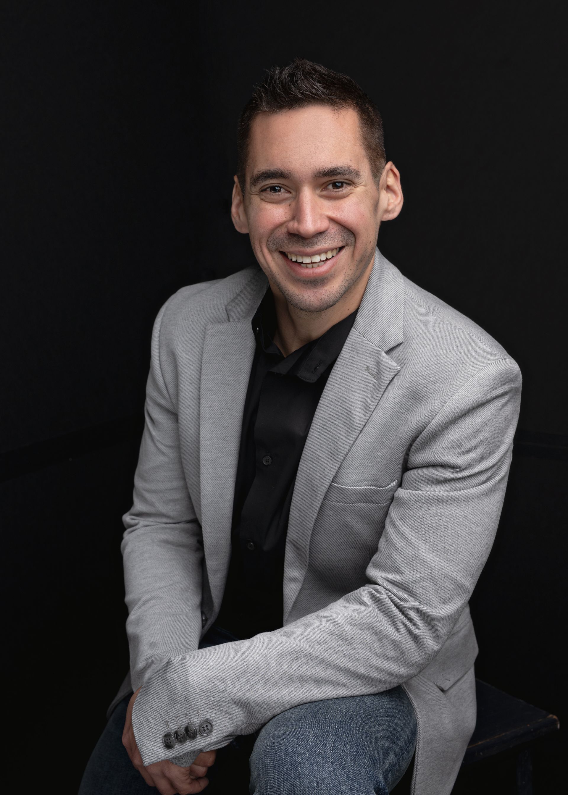 Man in blazer smiles at the camera, against a black background.