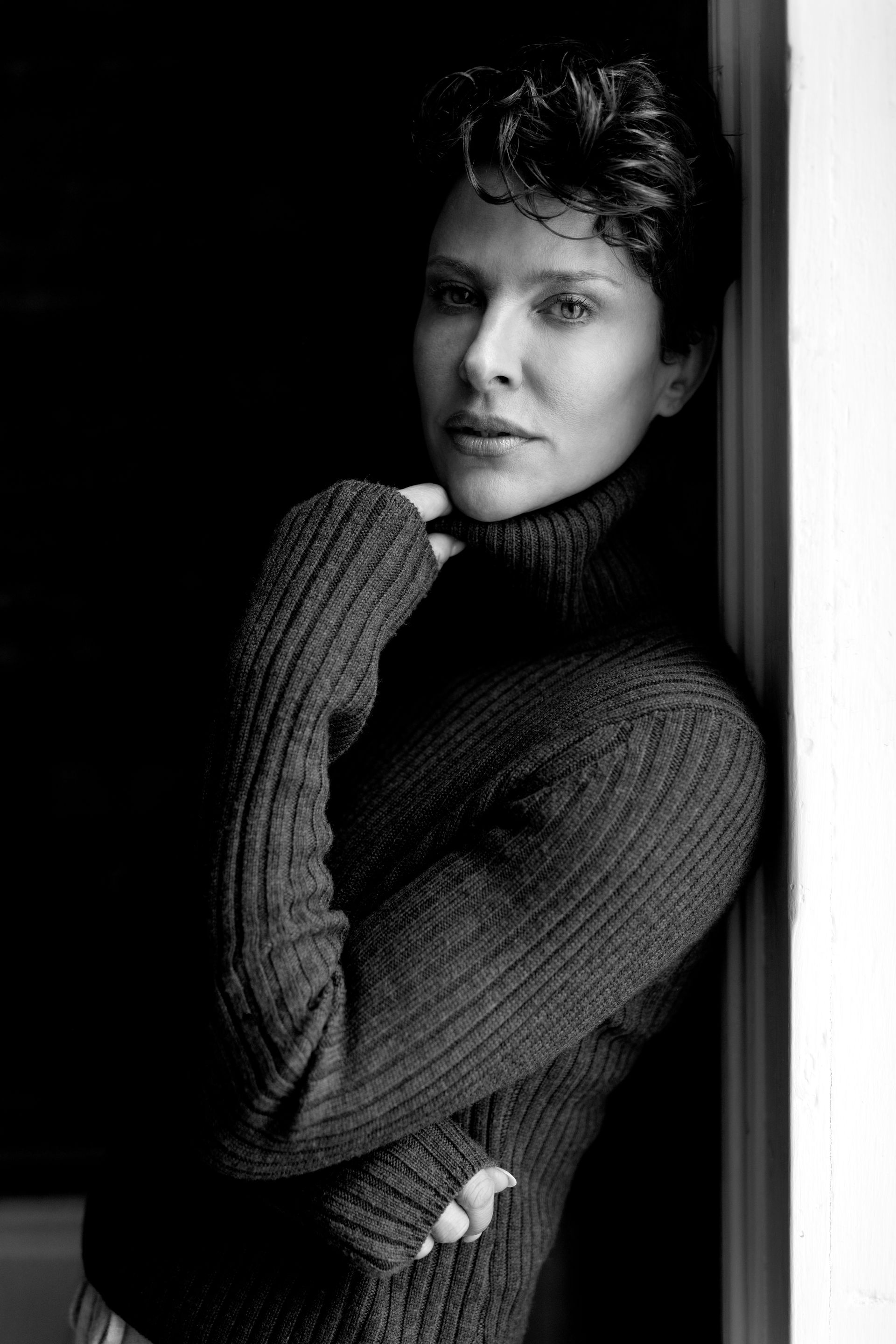 Woman in turtleneck sweater, leaning against a wall, looking at the camera, in black and white.