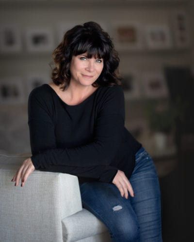 Woman with dark hair, leaning on a sofa, wearing black shirt and jeans, smiling.