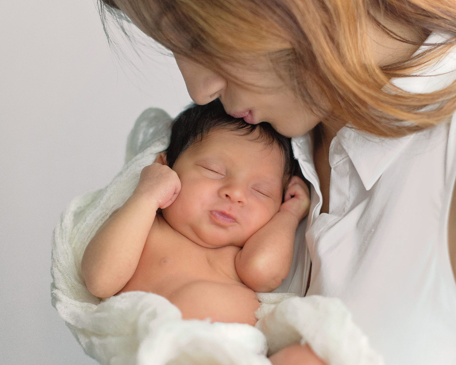 Woman kissing sleeping newborn, wrapped in a white blanket.