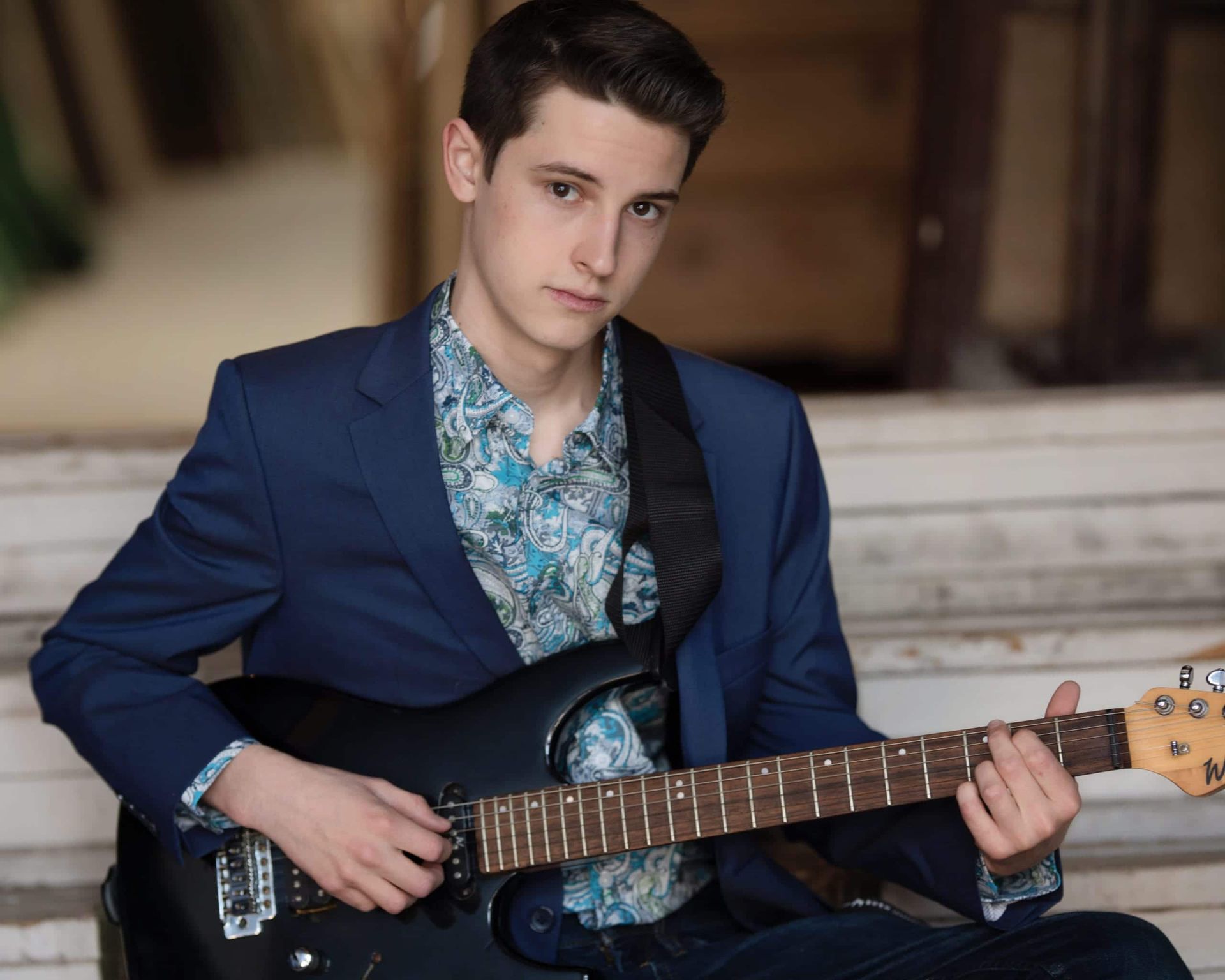 Young man in blue blazer and patterned shirt playing a black electric guitar, seated.