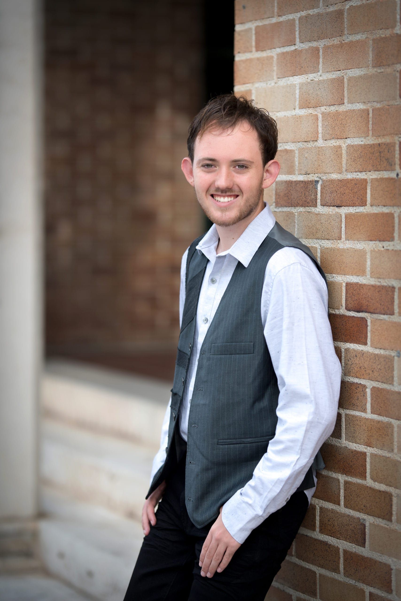 Man in a vest smiles while leaning against a brick wall outdoors.