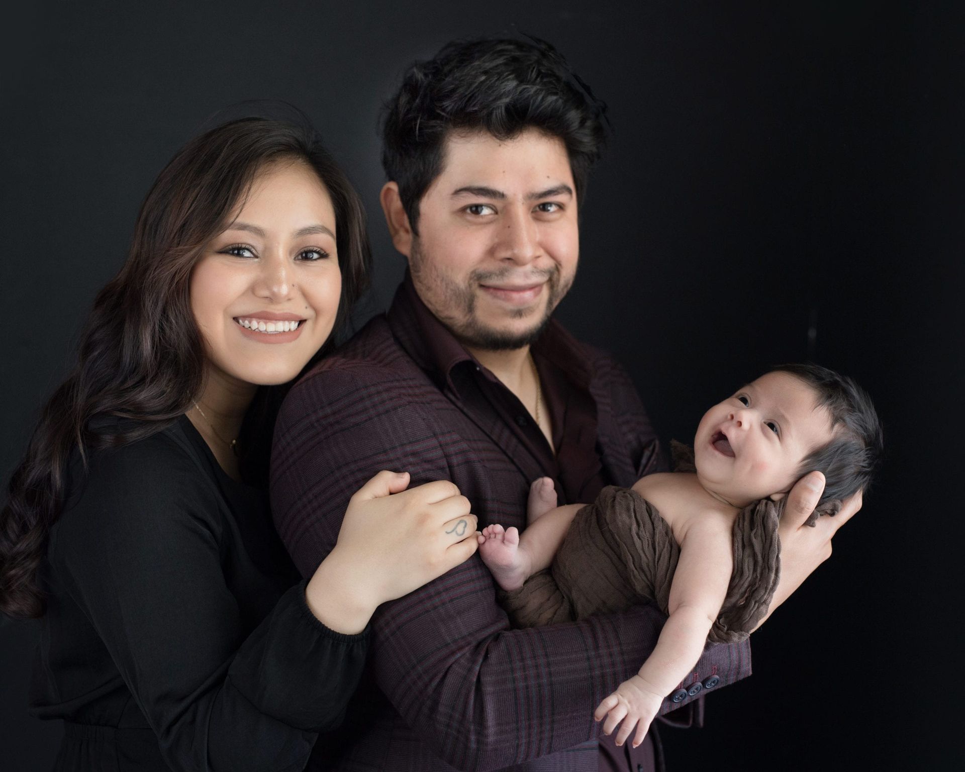 Parents holding newborn, smiling; dark background.