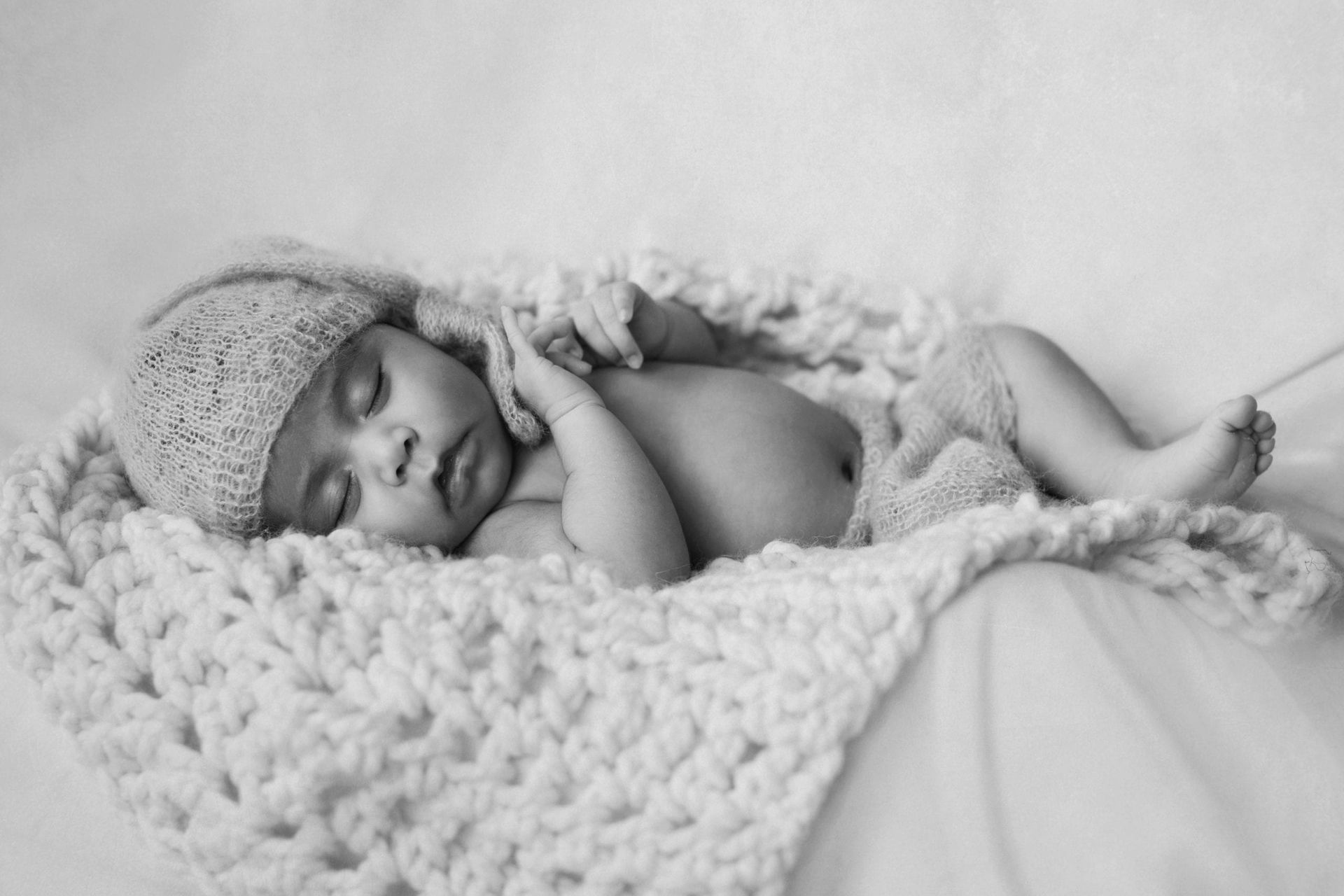 Sleeping newborn wrapped in a white, textured blanket, wearing a matching hat, on a white surface.