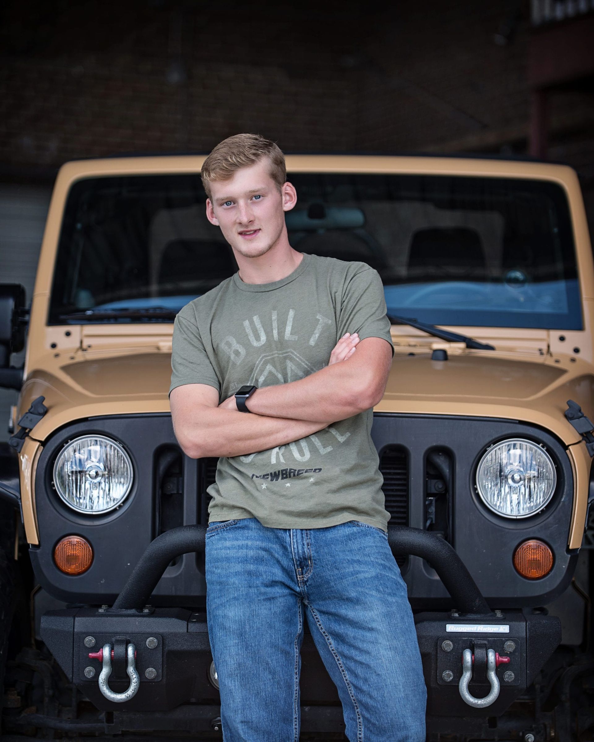 Man with arms crossed in front of tan Jeep. Wearing green shirt and jeans.