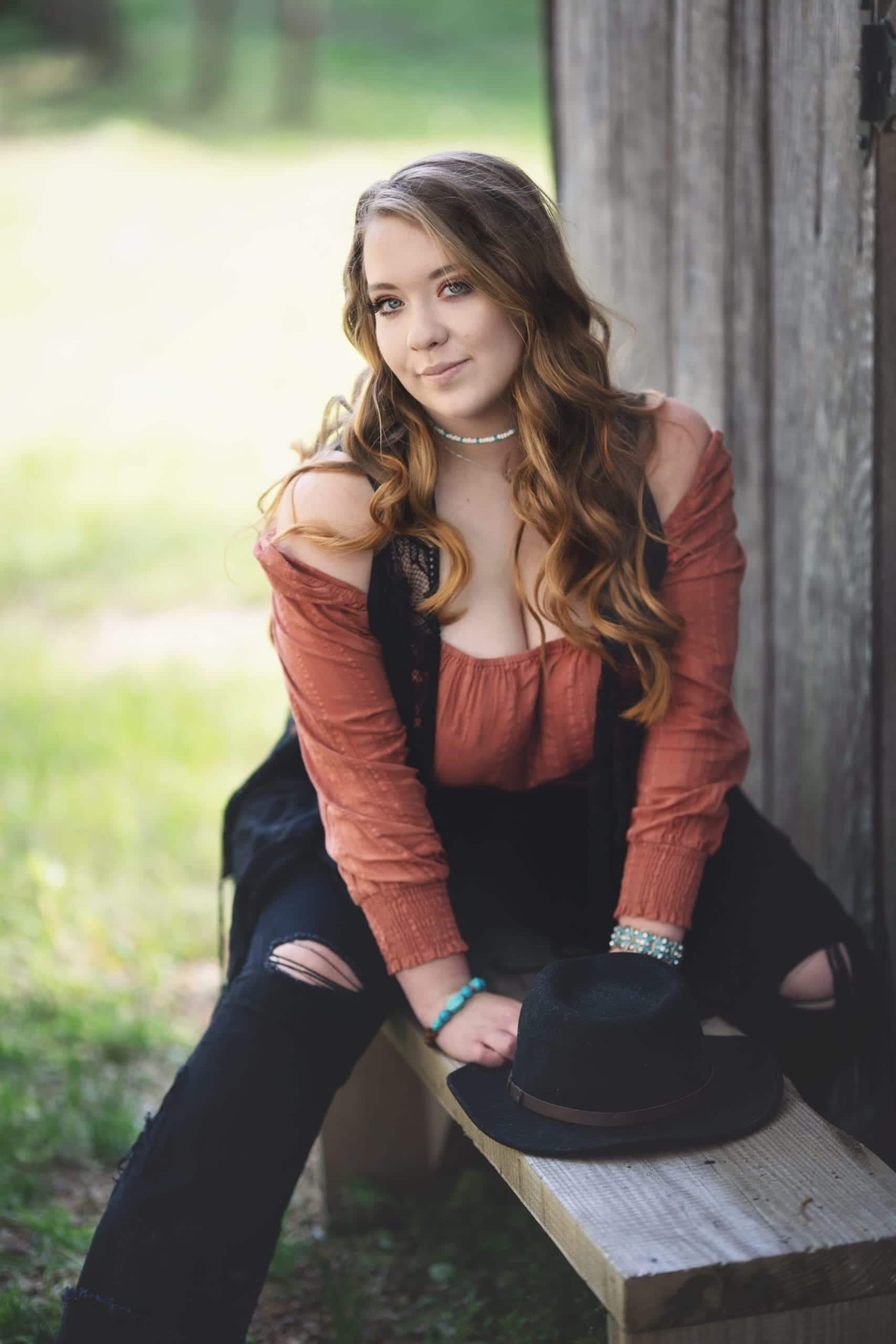 Woman with long wavy hair, smiling, sitting on a wooden bench, wearing a burnt orange shirt and ripped black jeans.
