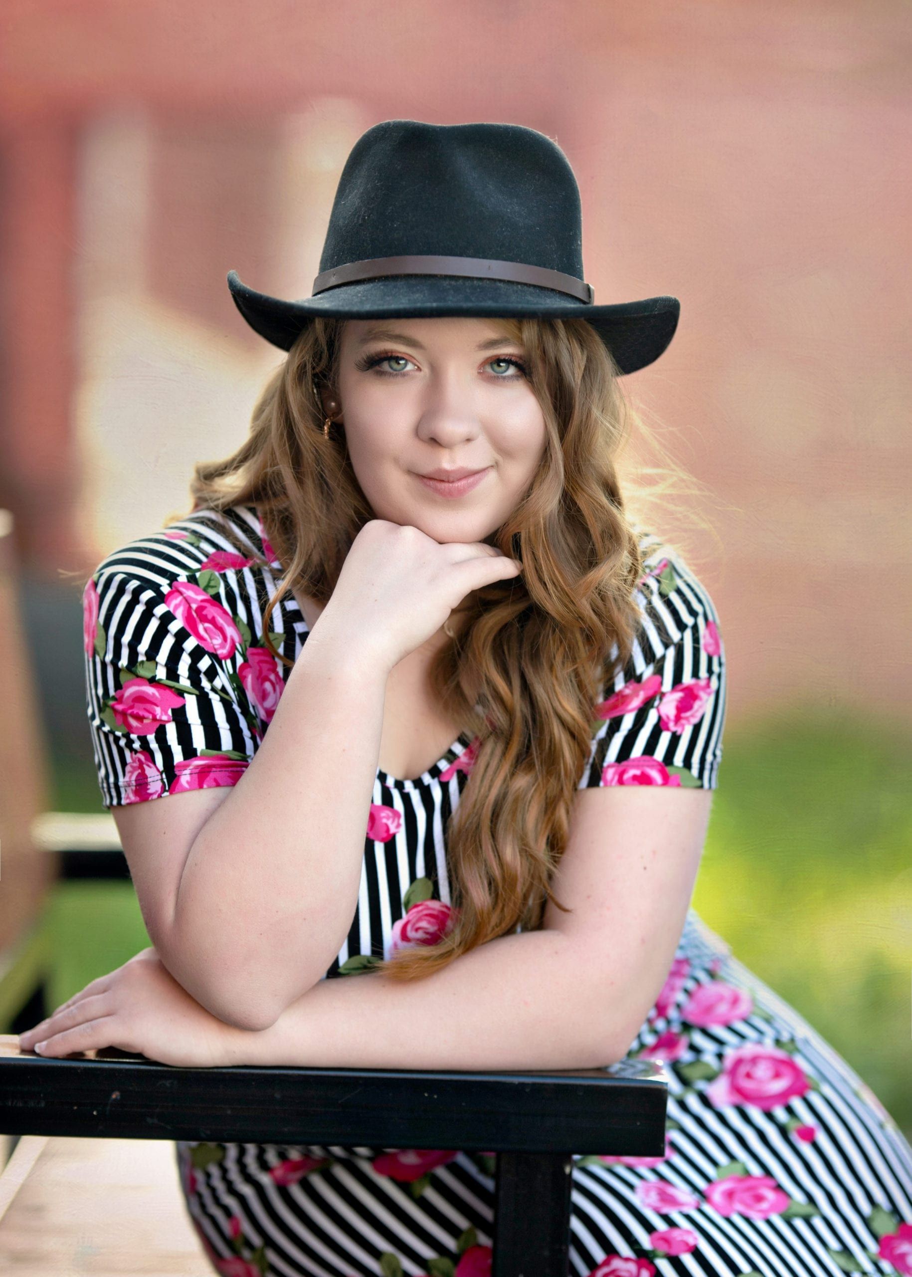 Woman wearing black hat and floral dress, leaning on a black surface, smiling, outdoors.
