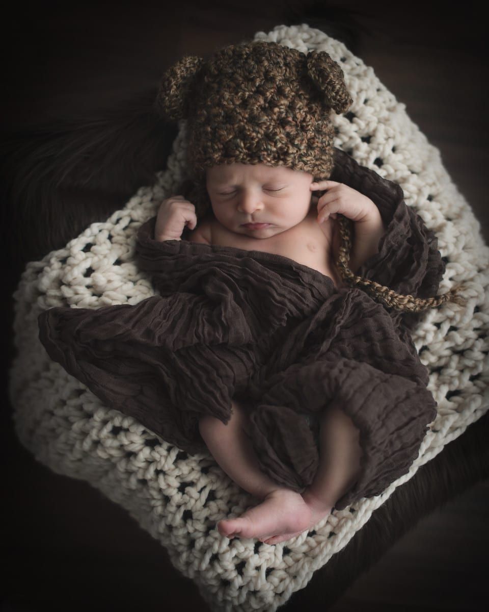 Newborn baby wearing a bear hat, wrapped in a brown blanket, lying on a cream-colored blanket.