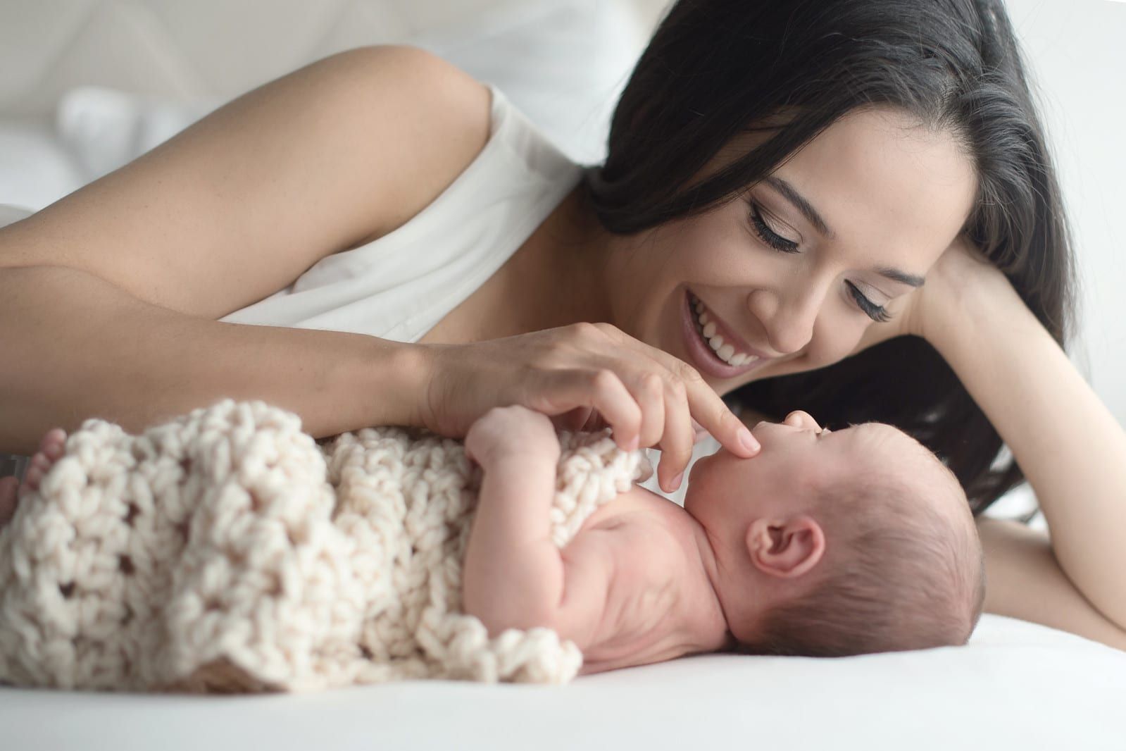 Mother smiles as she gently touches the face of her newborn, who is swaddled in a beige blanket.