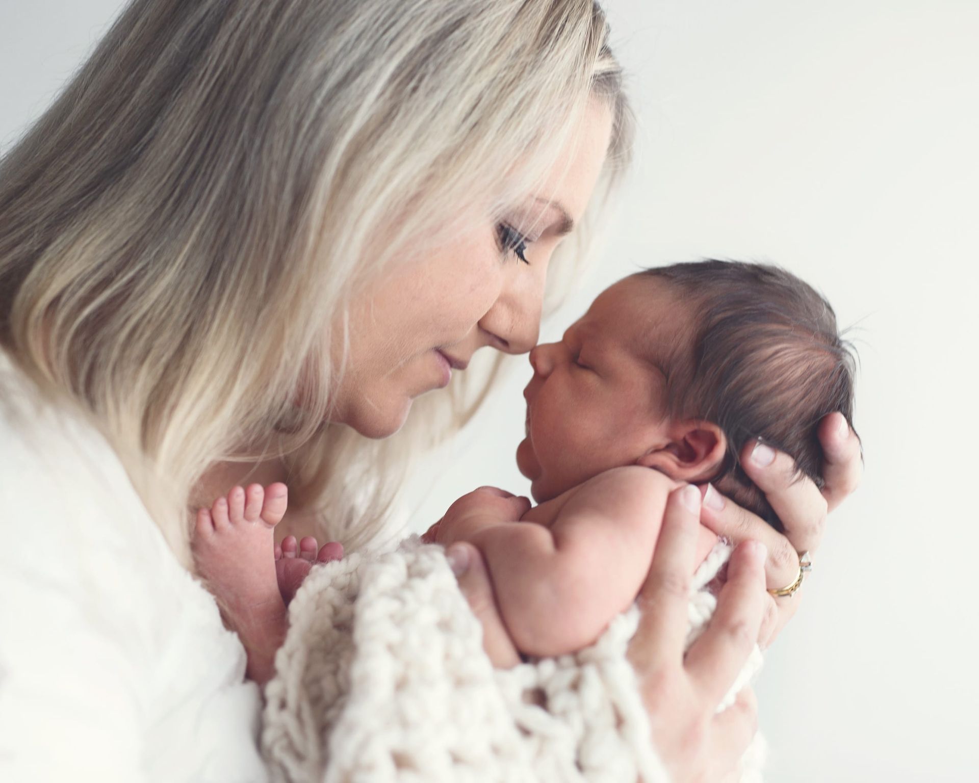 Woman with blonde hair lovingly holds a sleeping newborn baby wrapped in a cream-colored blanket.