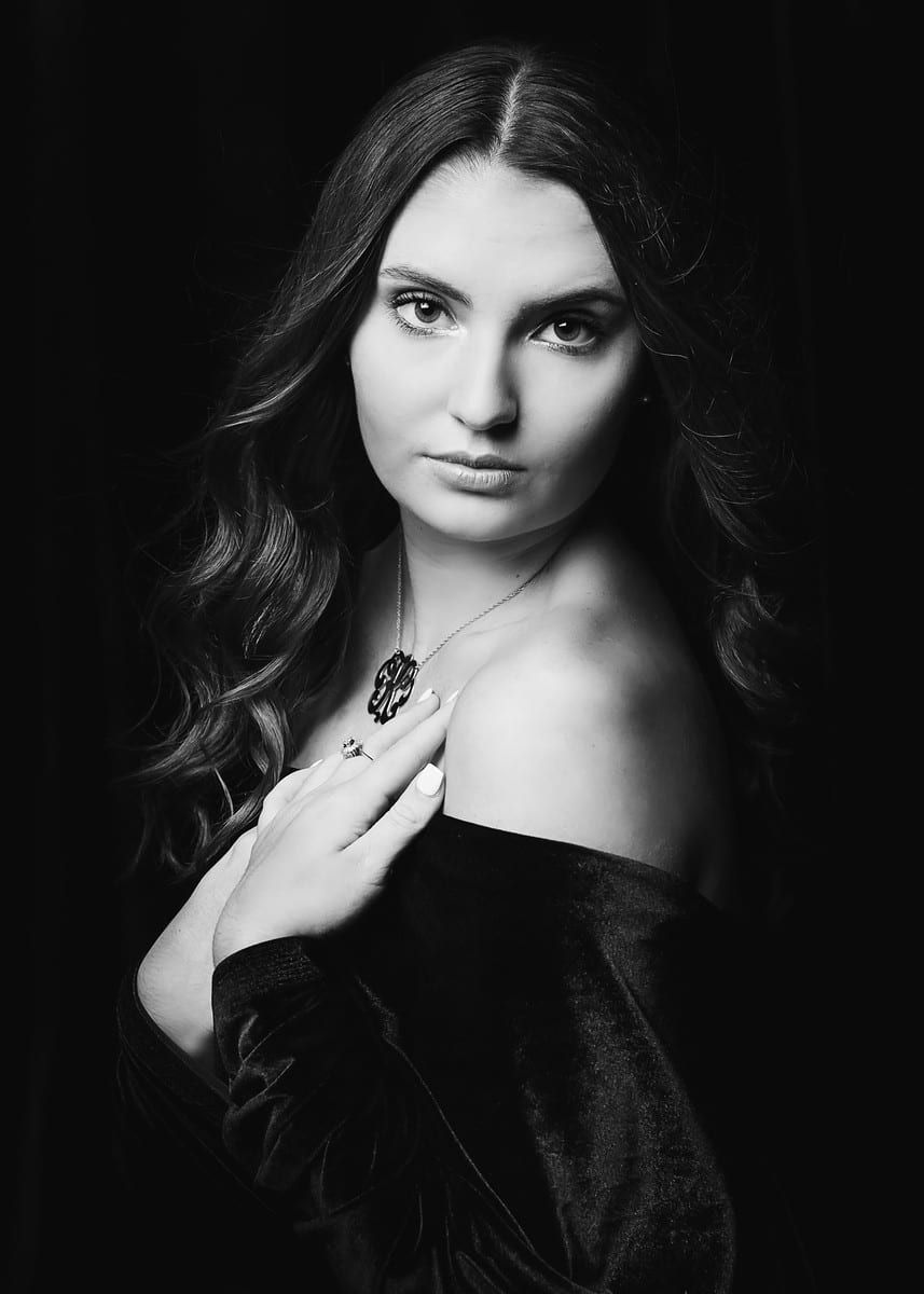 Woman with dark hair and a necklace, looking at the camera, wearing a black off-the-shoulder top.