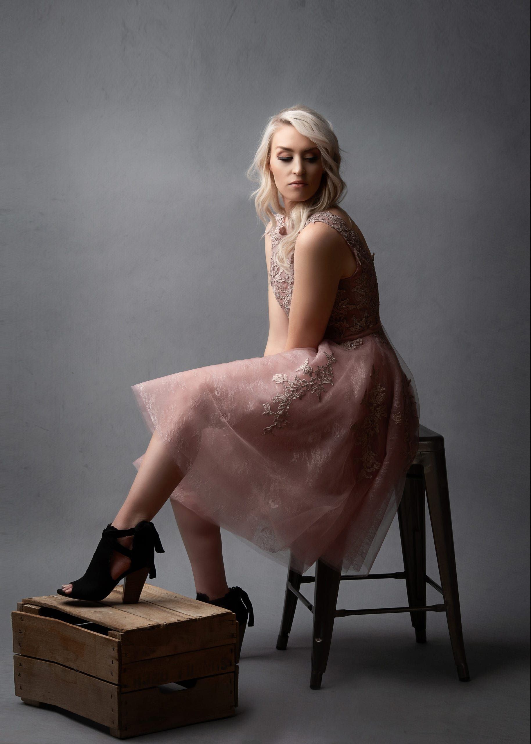 Woman in pink dress, blonde hair, posing on stool and wooden box; gray background.