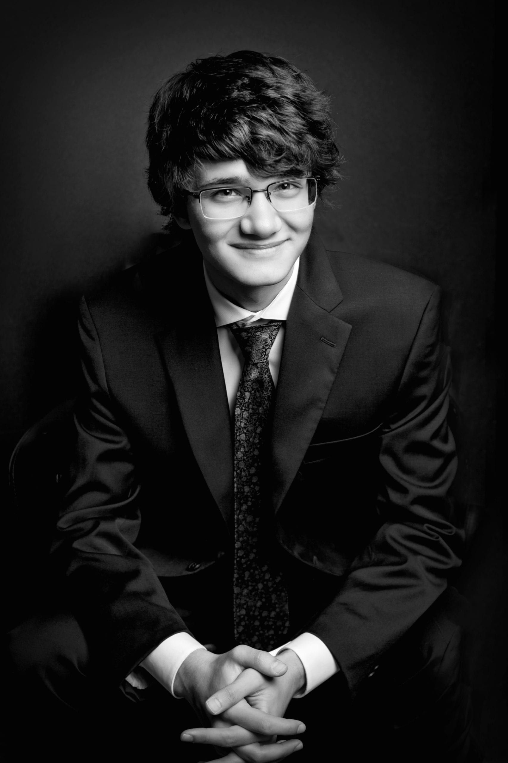 Young man in suit and glasses, smiling, hands clasped, in a formal studio setting.