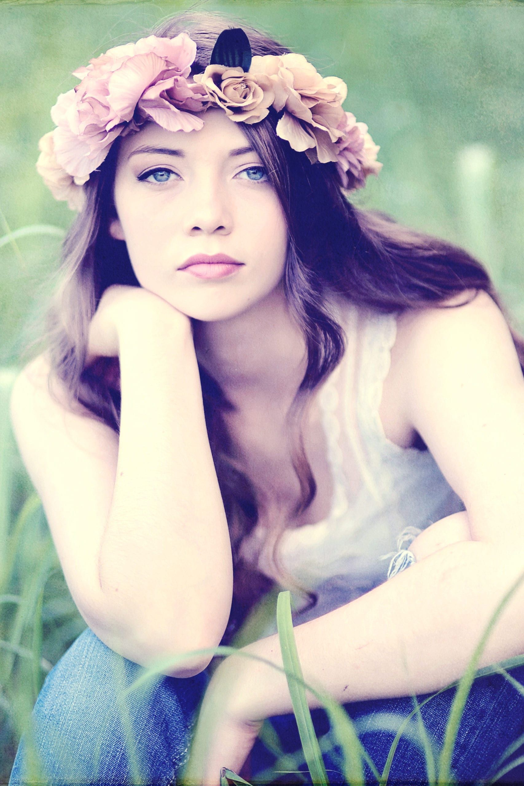 Woman with flower crown, sitting in grass, resting head on hand, blue eyes, looking at camera.