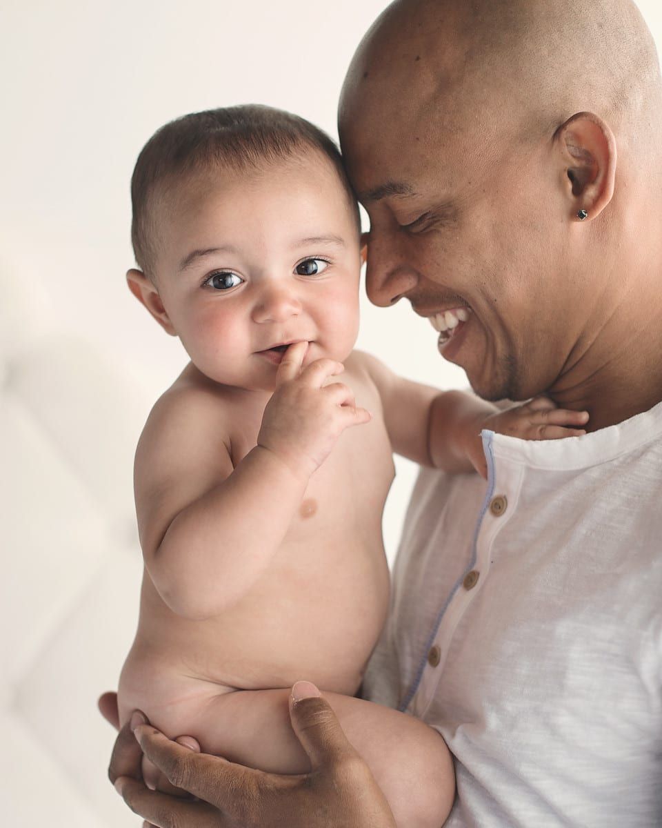 Father smiling, holding baby. Baby is smiling, fingers in mouth. Light skin, soft background.