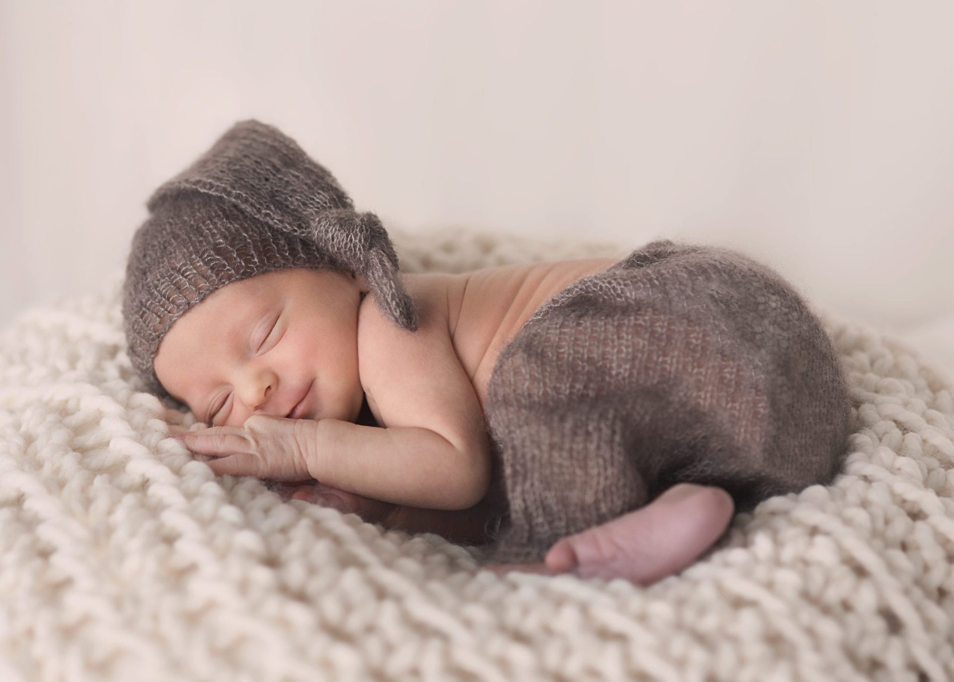 Baby asleep on a white blanket wearing a brown hat and pants.