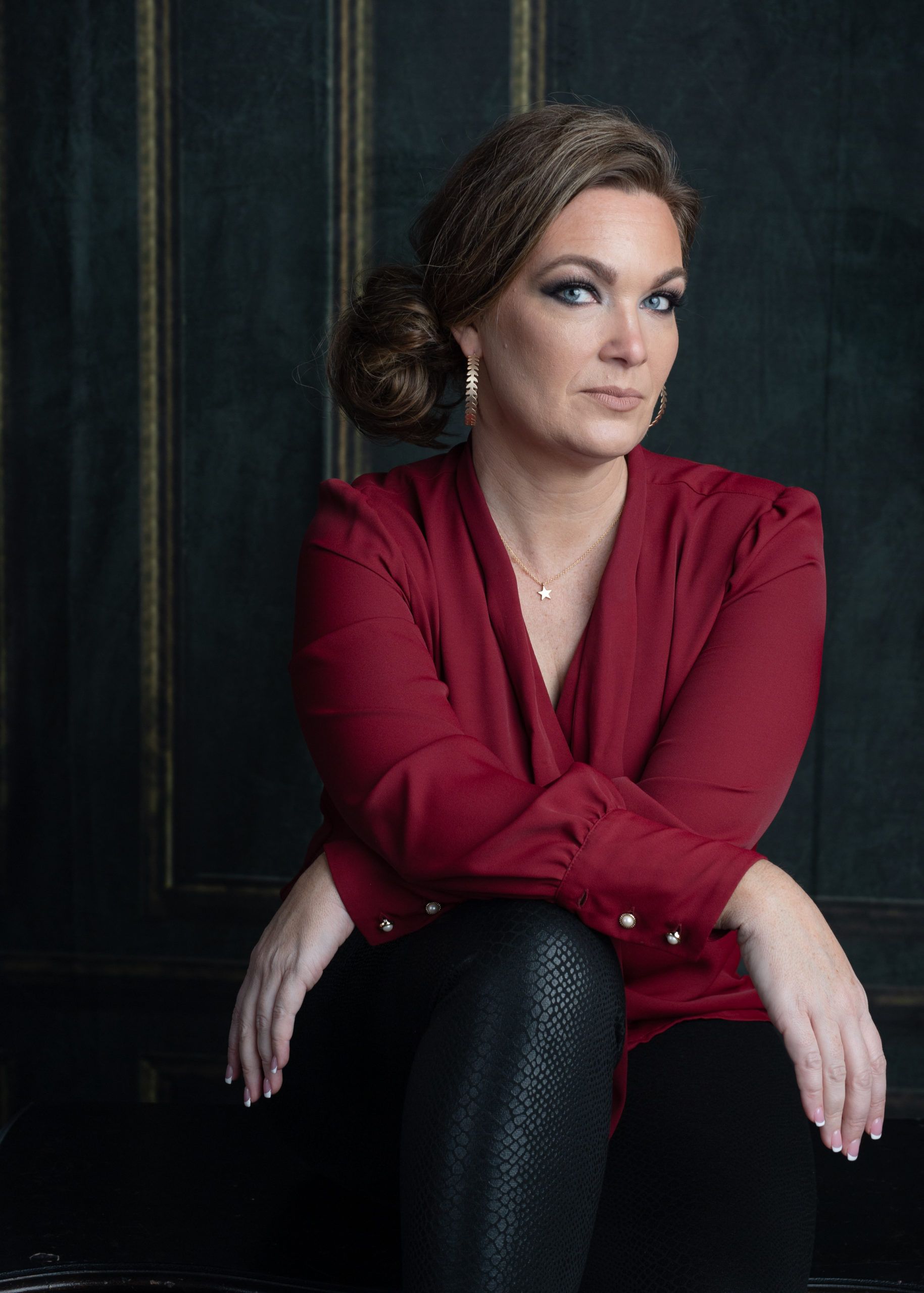 Woman with dark hair, in red blouse and black pants, seated with arms crossed, looking at camera.