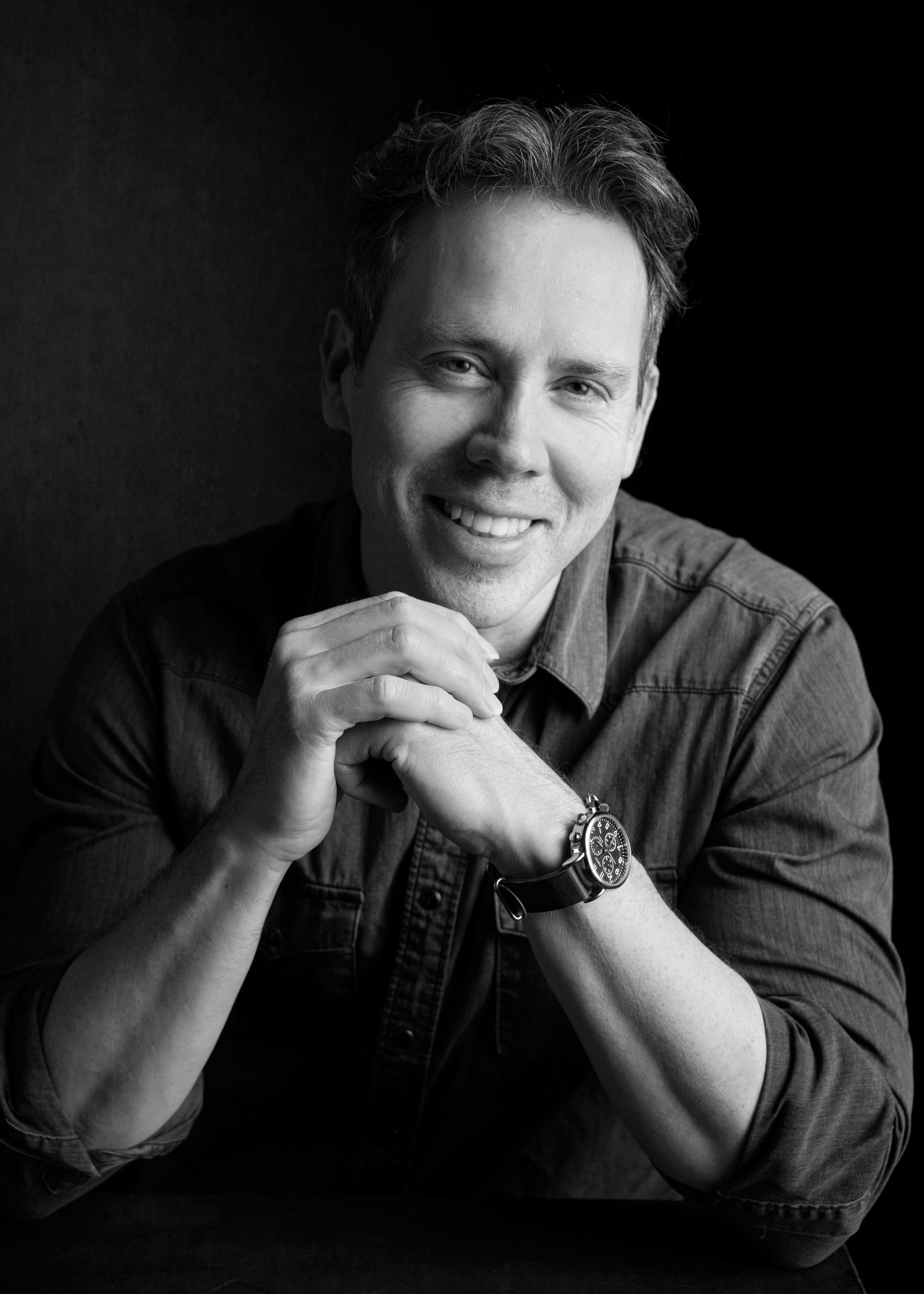 Man with short hair smiles, wearing a dark shirt and a watch, with hands clasped.