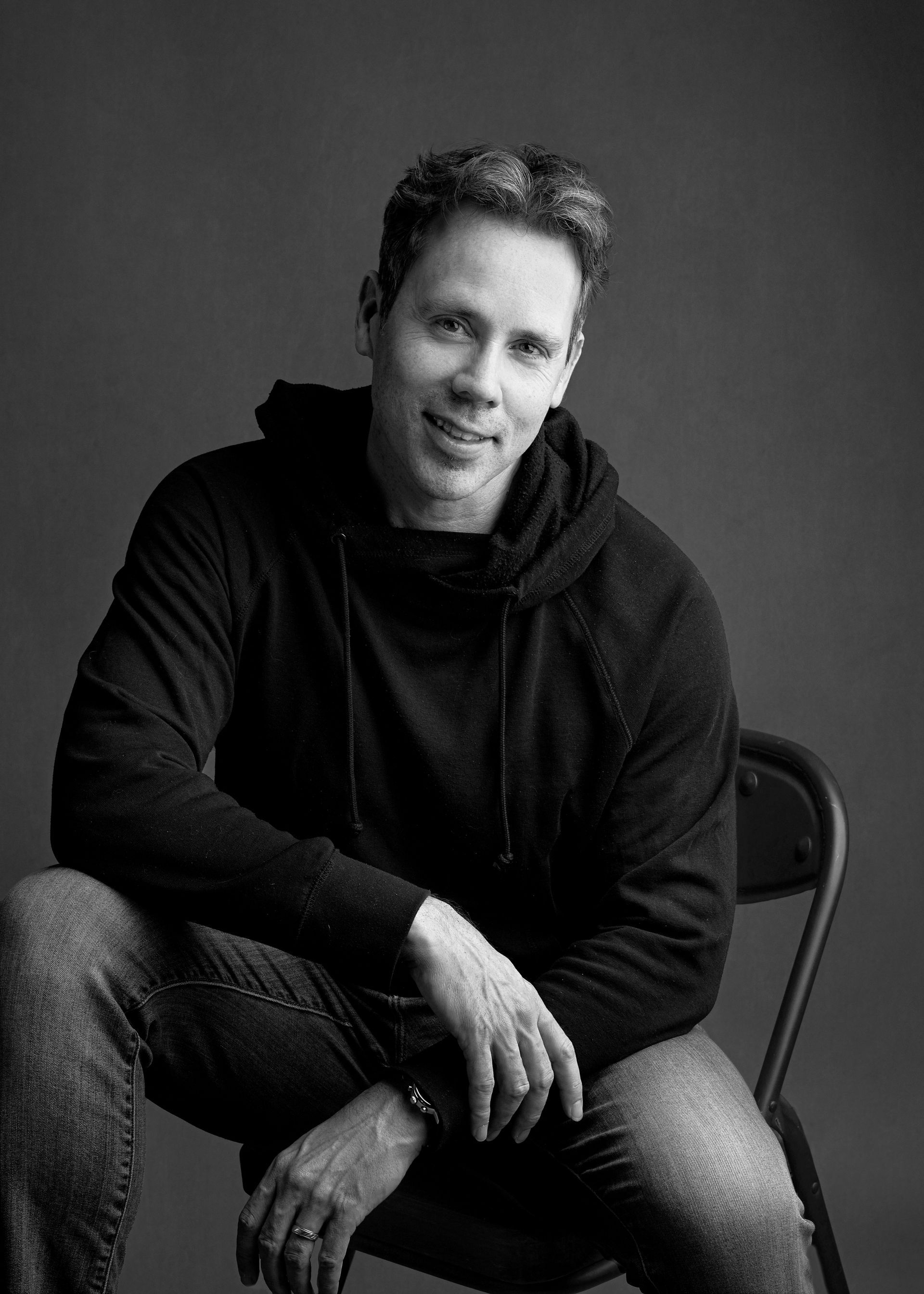 Man in hoodie and jeans, sitting on a chair, smiling. Black and white photo.