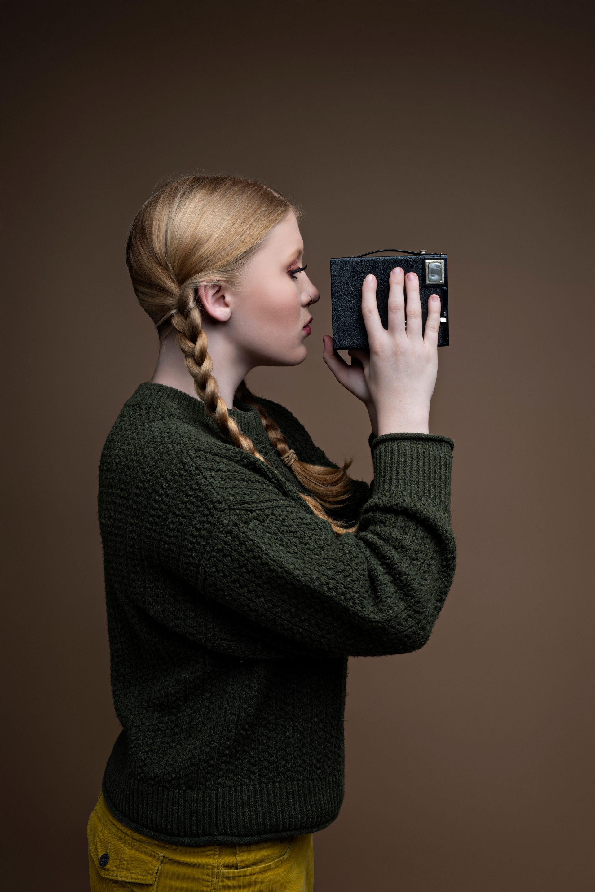 Blonde woman in green sweater takes photo with a black box camera; brown backdrop.