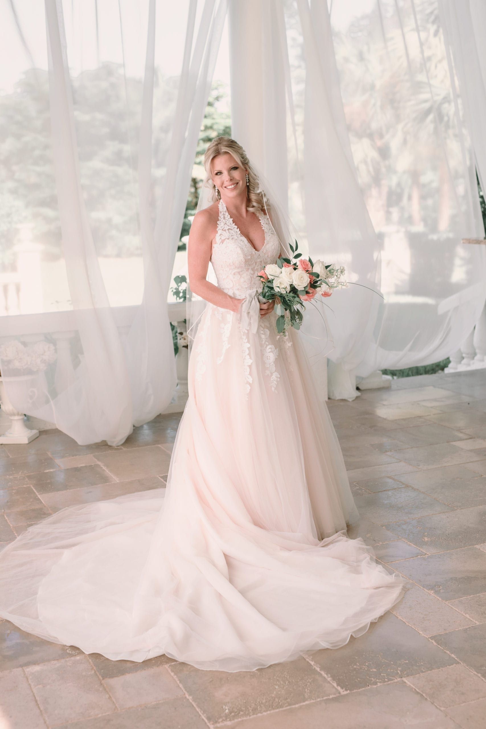 Blonde bride in a white and blush gown holds a bouquet. She's smiling in a bright, open-air space.