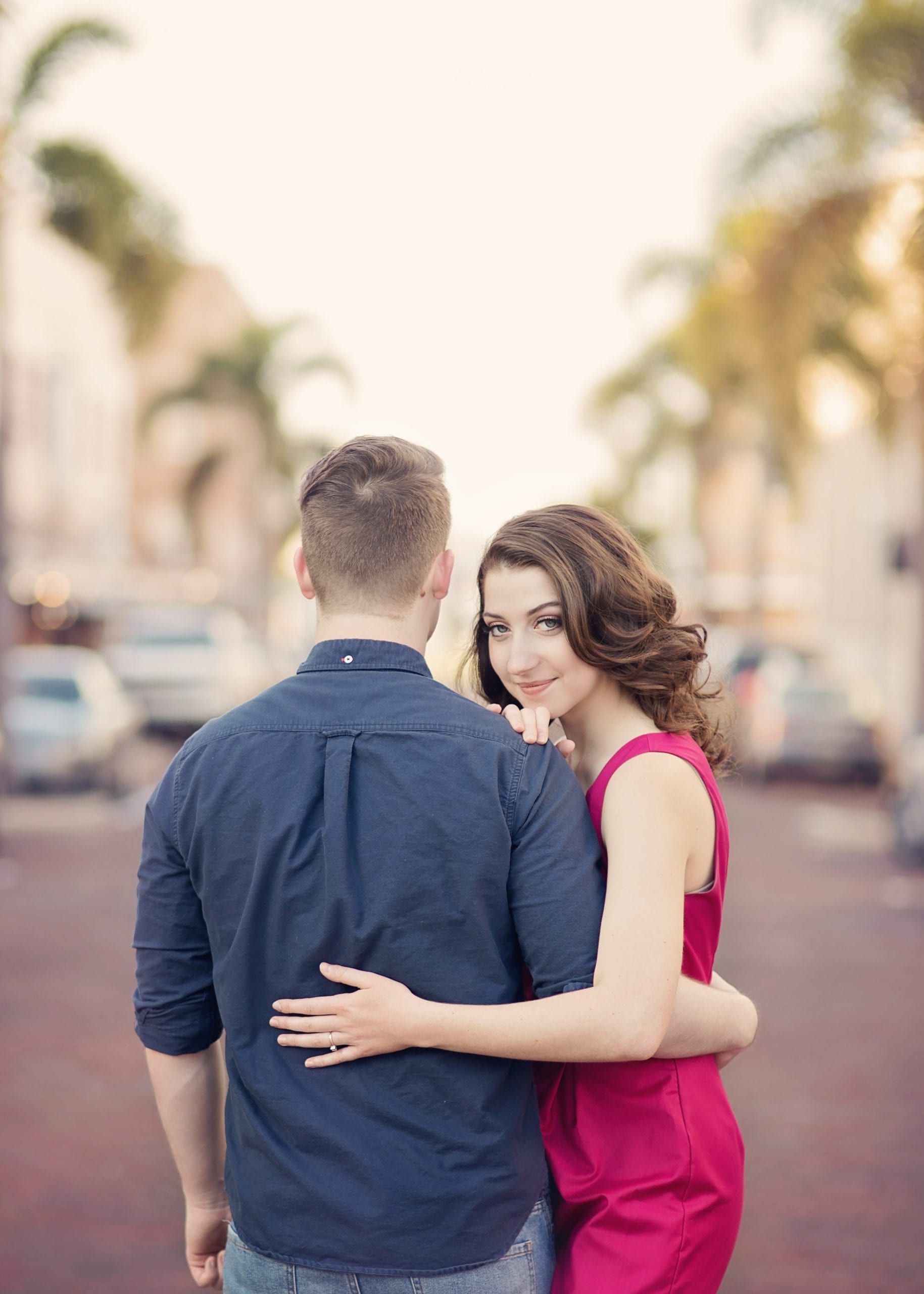 Woman in red dress hugs man in blue shirt, looking over shoulder, on a brick street.