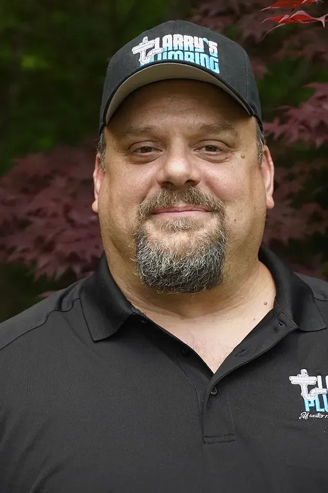 Man with a goatee wearing a black hat and shirt with a plumbing logo.