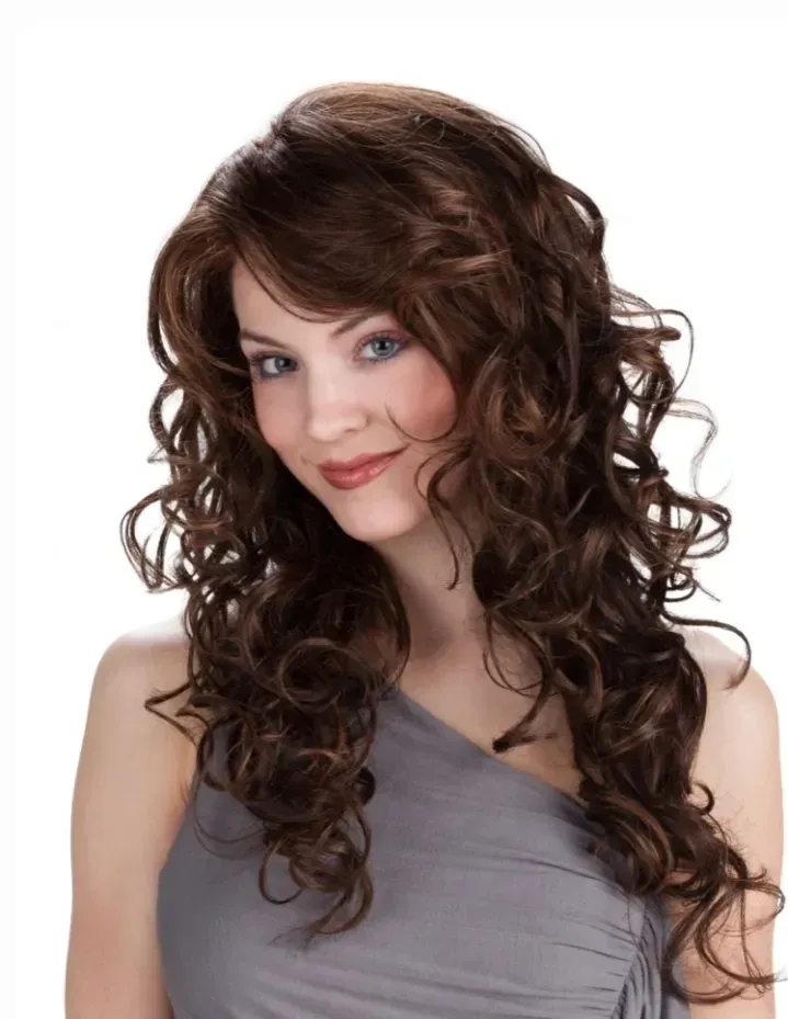 Woman with long curly brown hair wearing a gray one-shoulder top on a white background