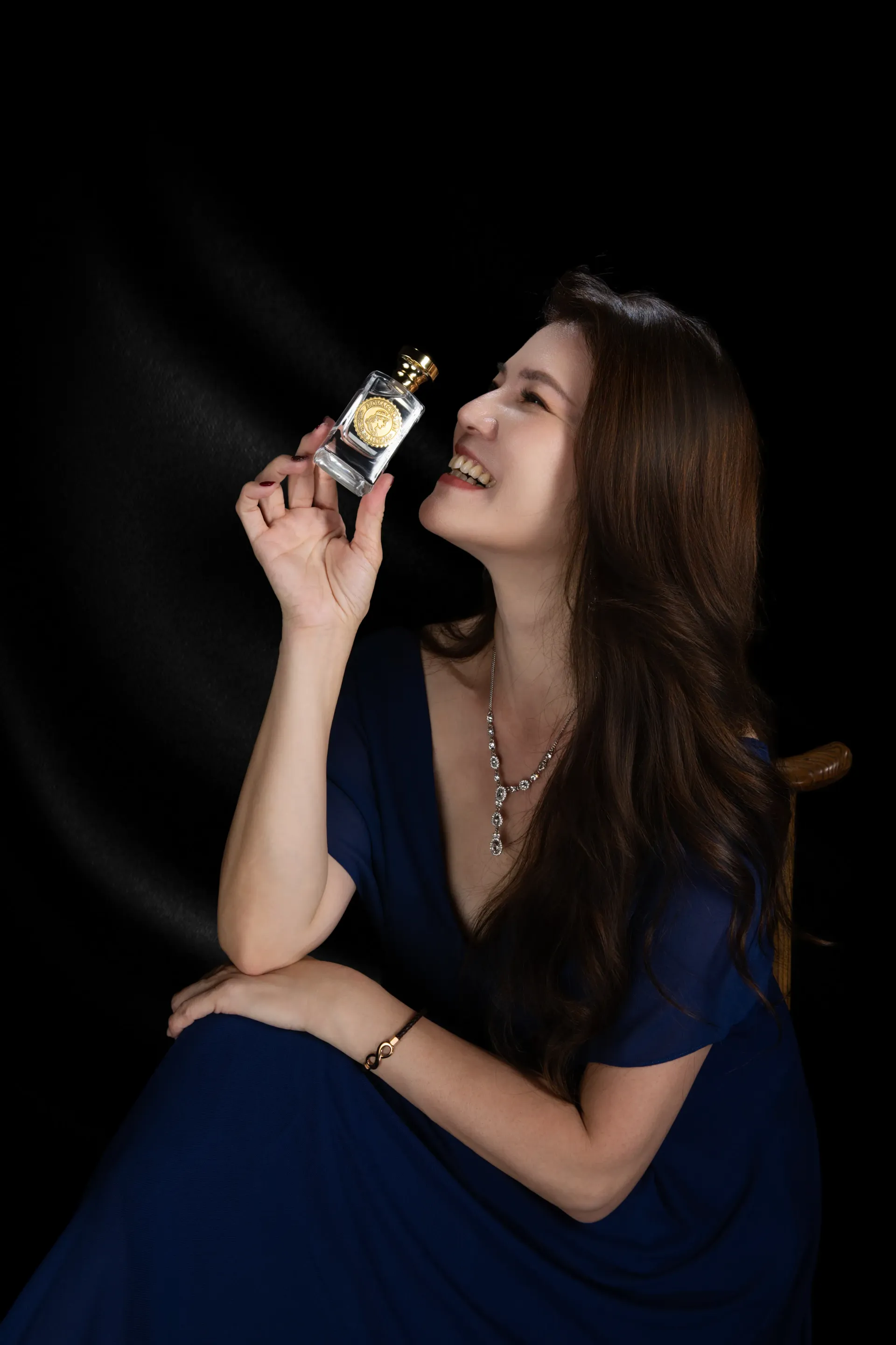 身穿藍色洋裝的女子手持香水，面帶微笑，抬頭望向天空，背景為黑色。