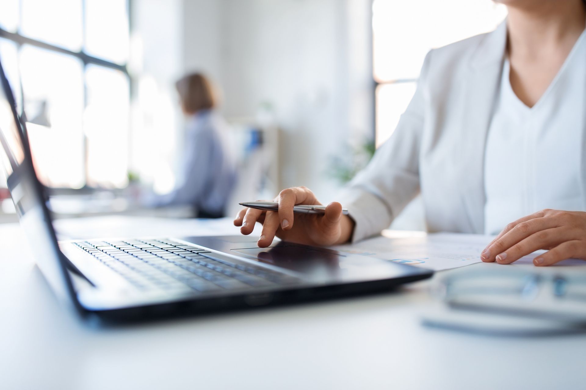 Woman in white blazer working on a laptop at a desk in an office.