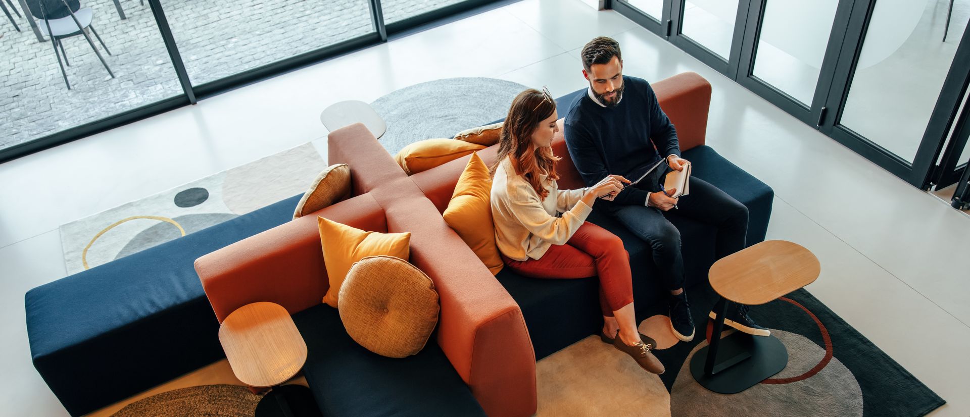 Two people on a modern sofa, looking at a tablet. They are in a bright, modern lobby.