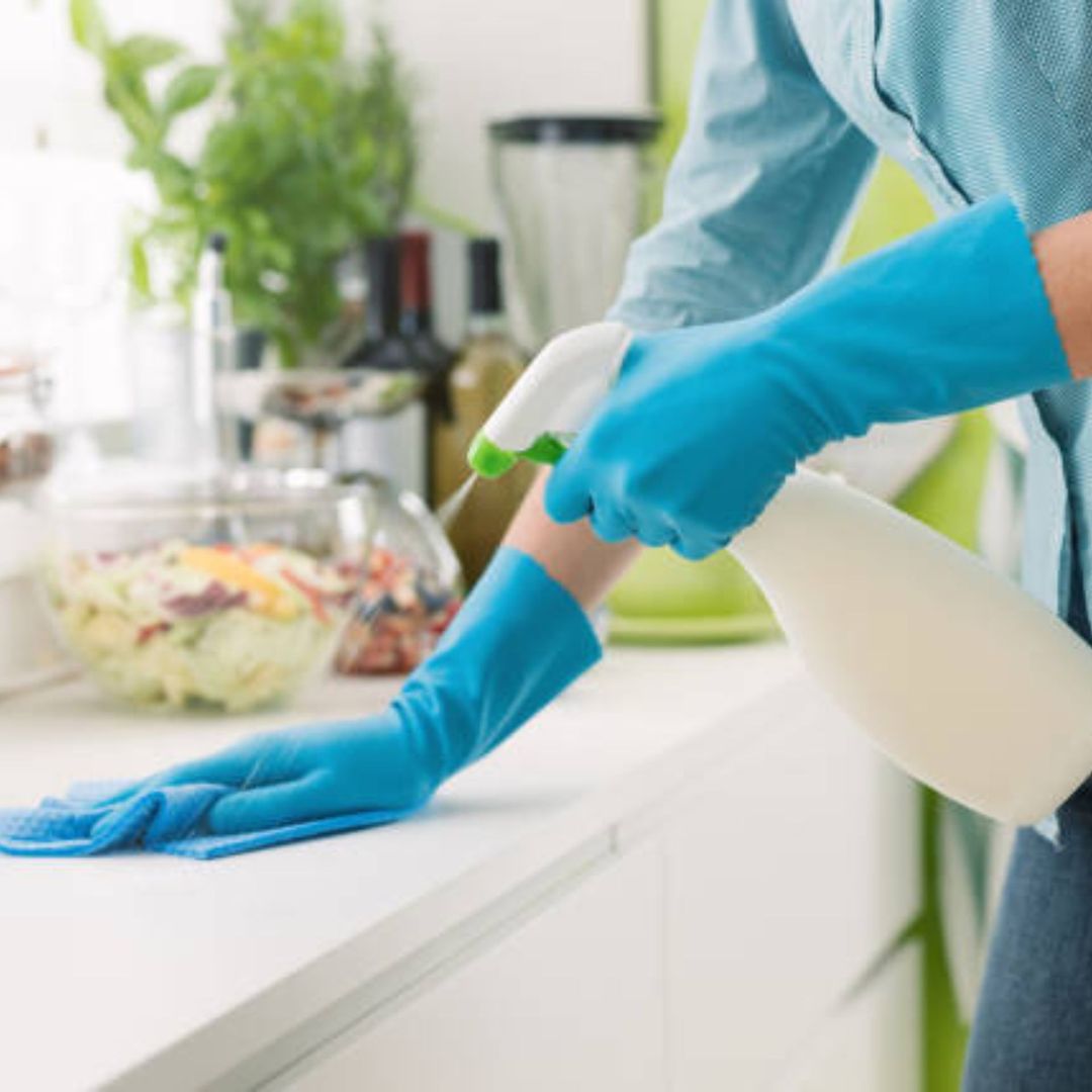 Person wearing blue gloves, cleaning a white kitchen countertop with spray bottle and cloth.